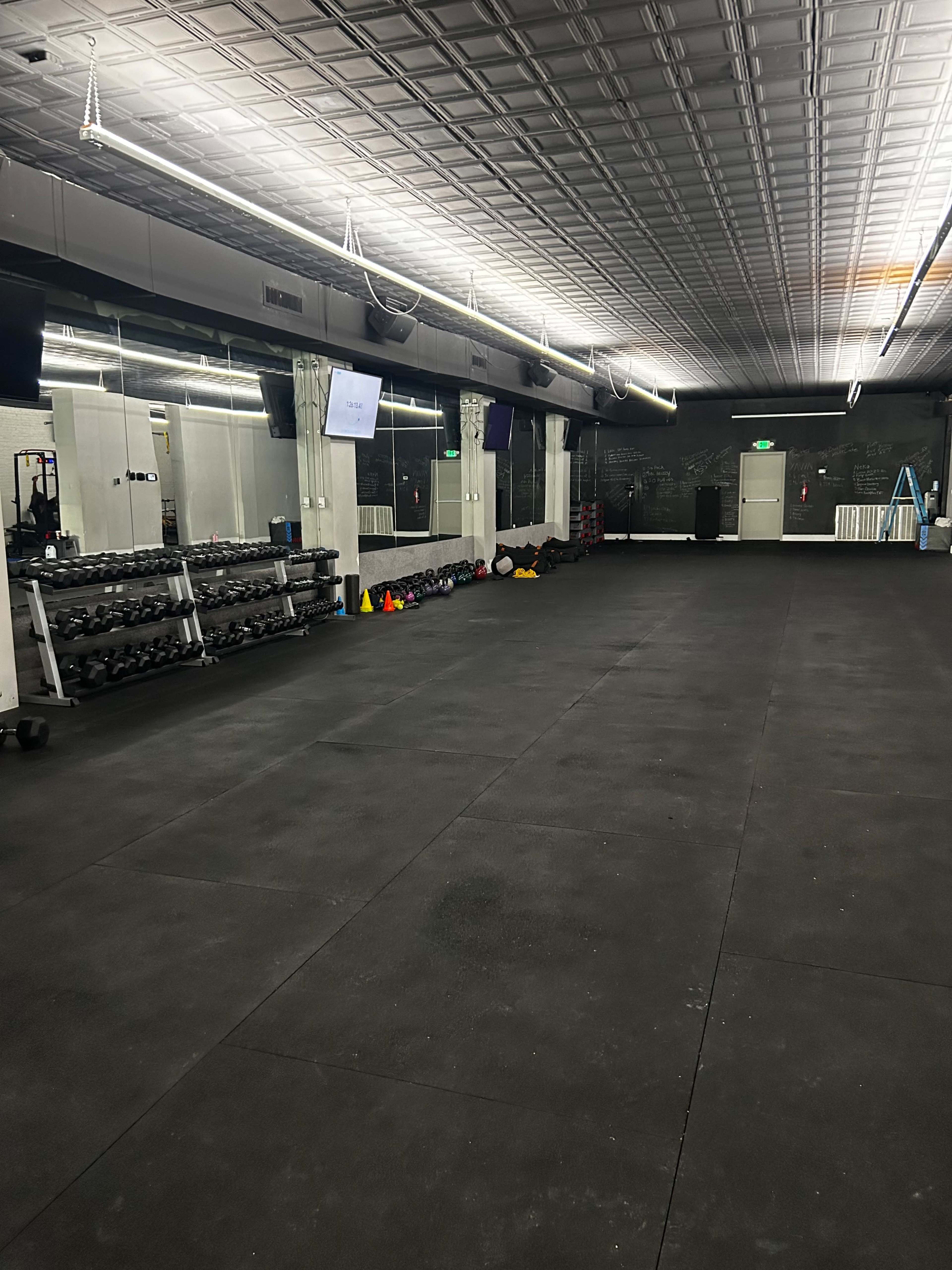 The image shows an empty gym space with rubber flooring, dumbbells neatly stacked along one wall, and a few exercise props scattered across the floor.