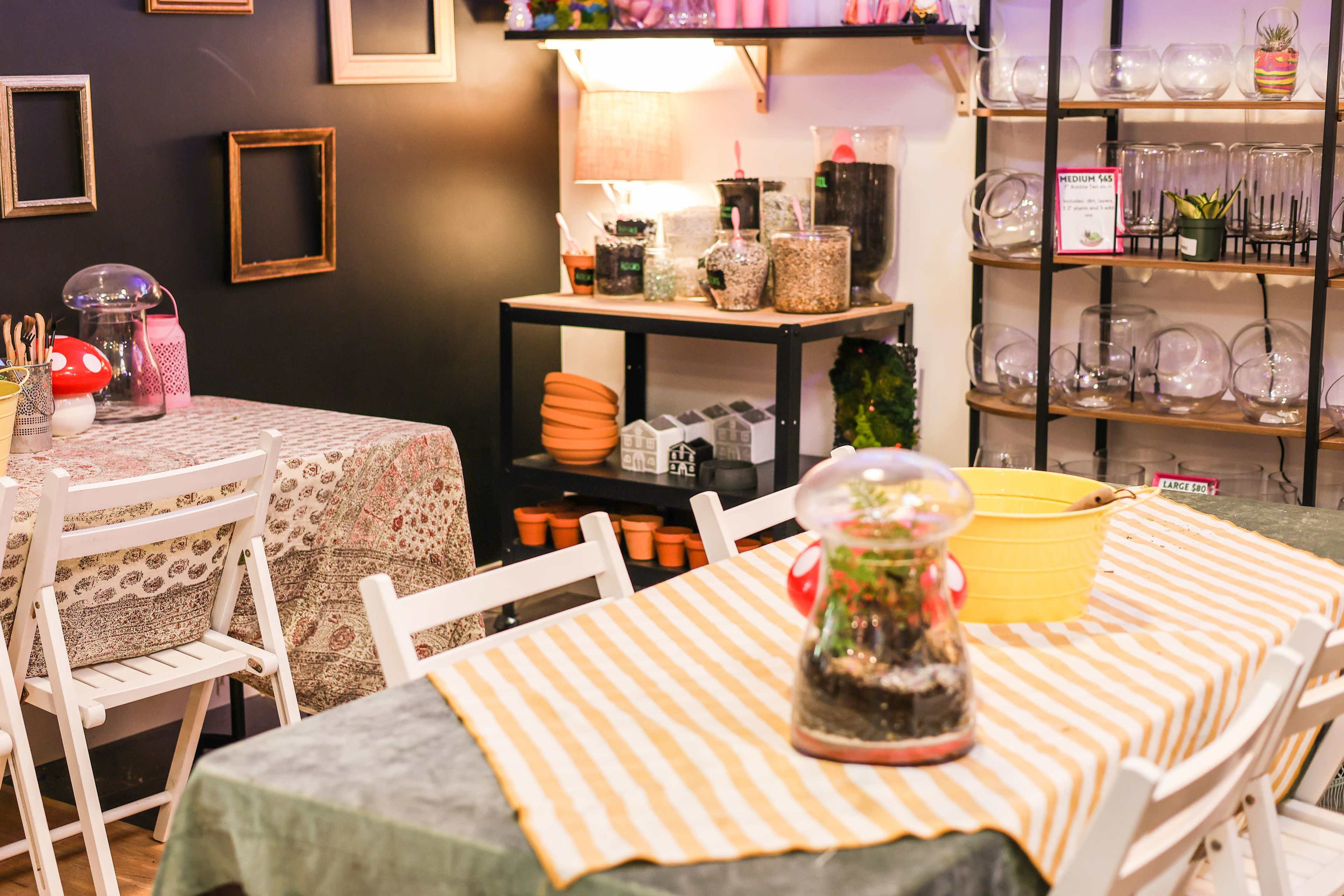A cozy interior of a café or workshop with tables covered in patterned cloths and shelves stocked with various gardening supplies and glass containers.