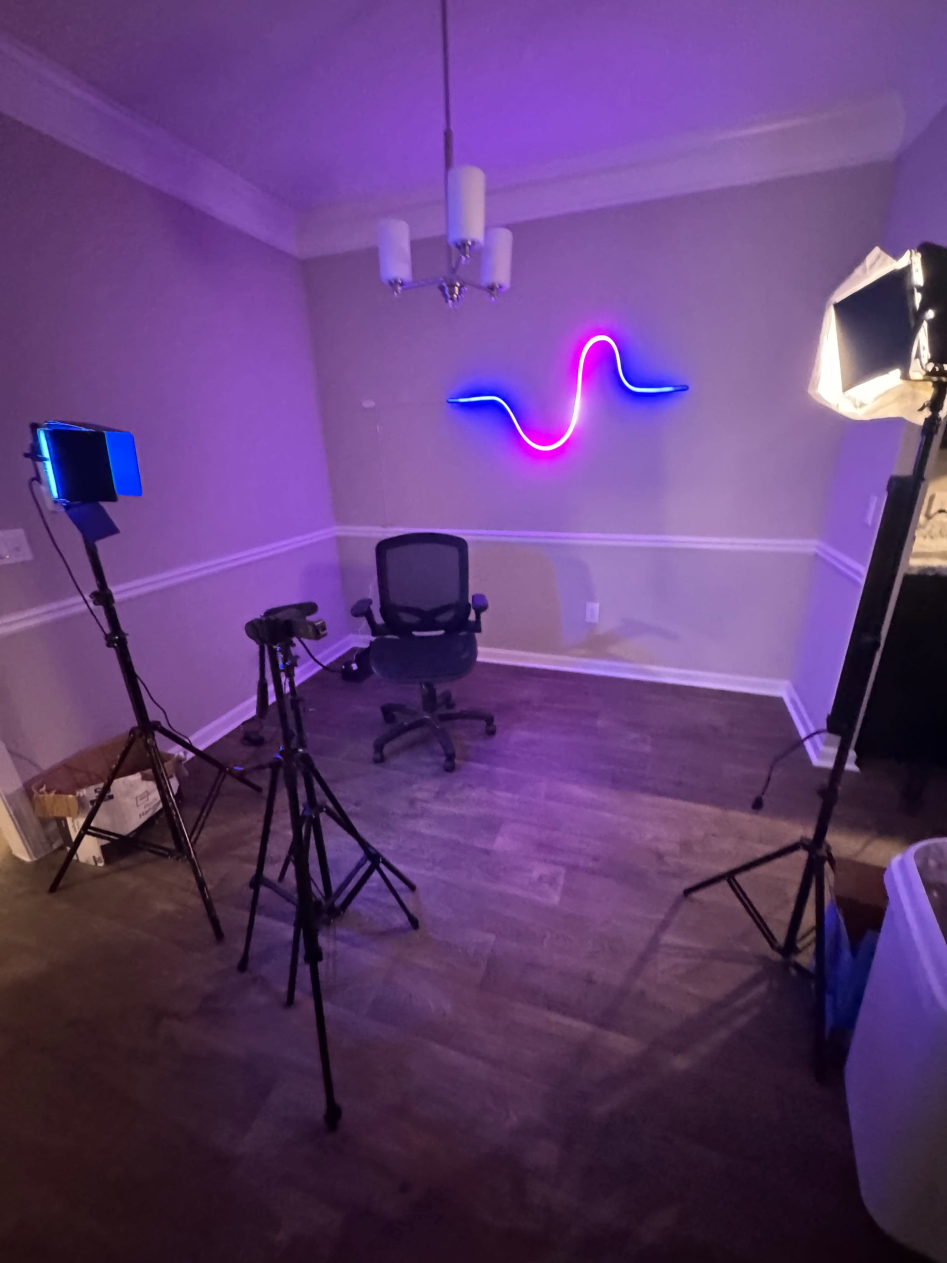 The image shows a dimly lit room with a black chair in the center, flanked by two lighting setups and a neon wave light on the wall.