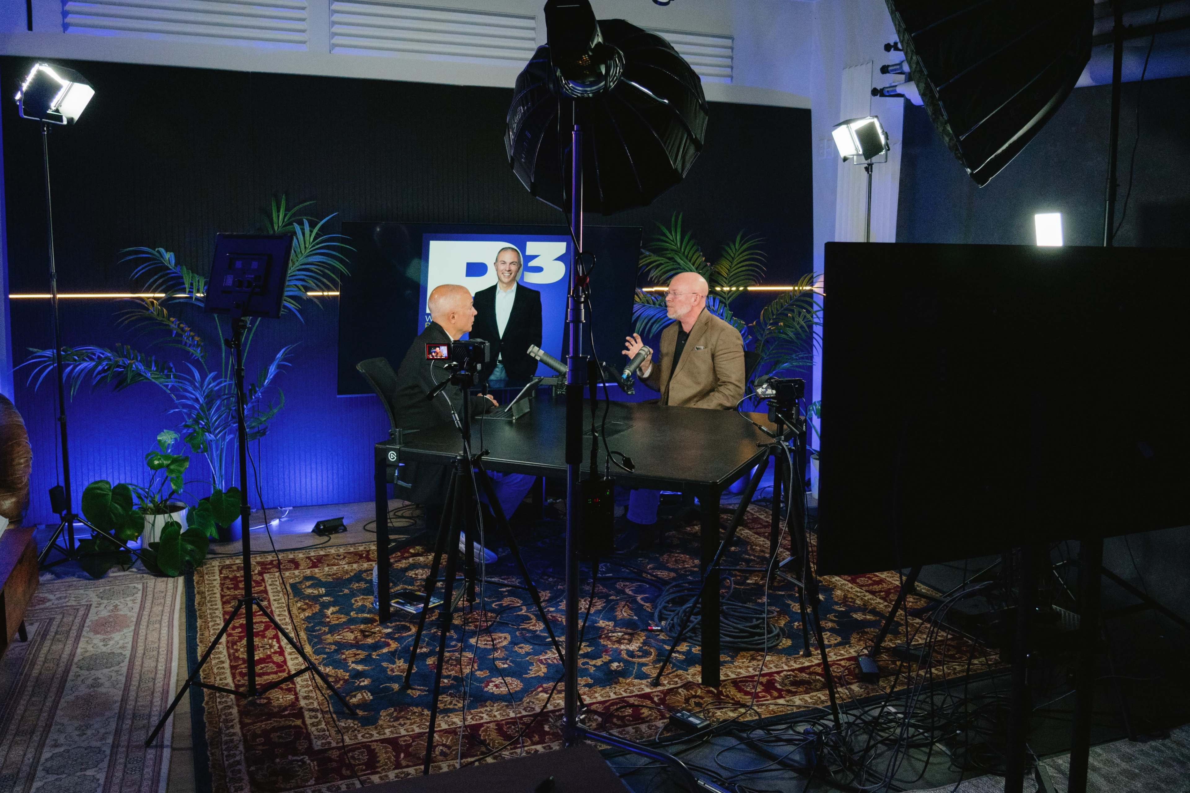 Two men are seated at a table in a well-lit studio, engaged in conversation, while a third man stands behind them in front of a large screen displaying the letters "P3."