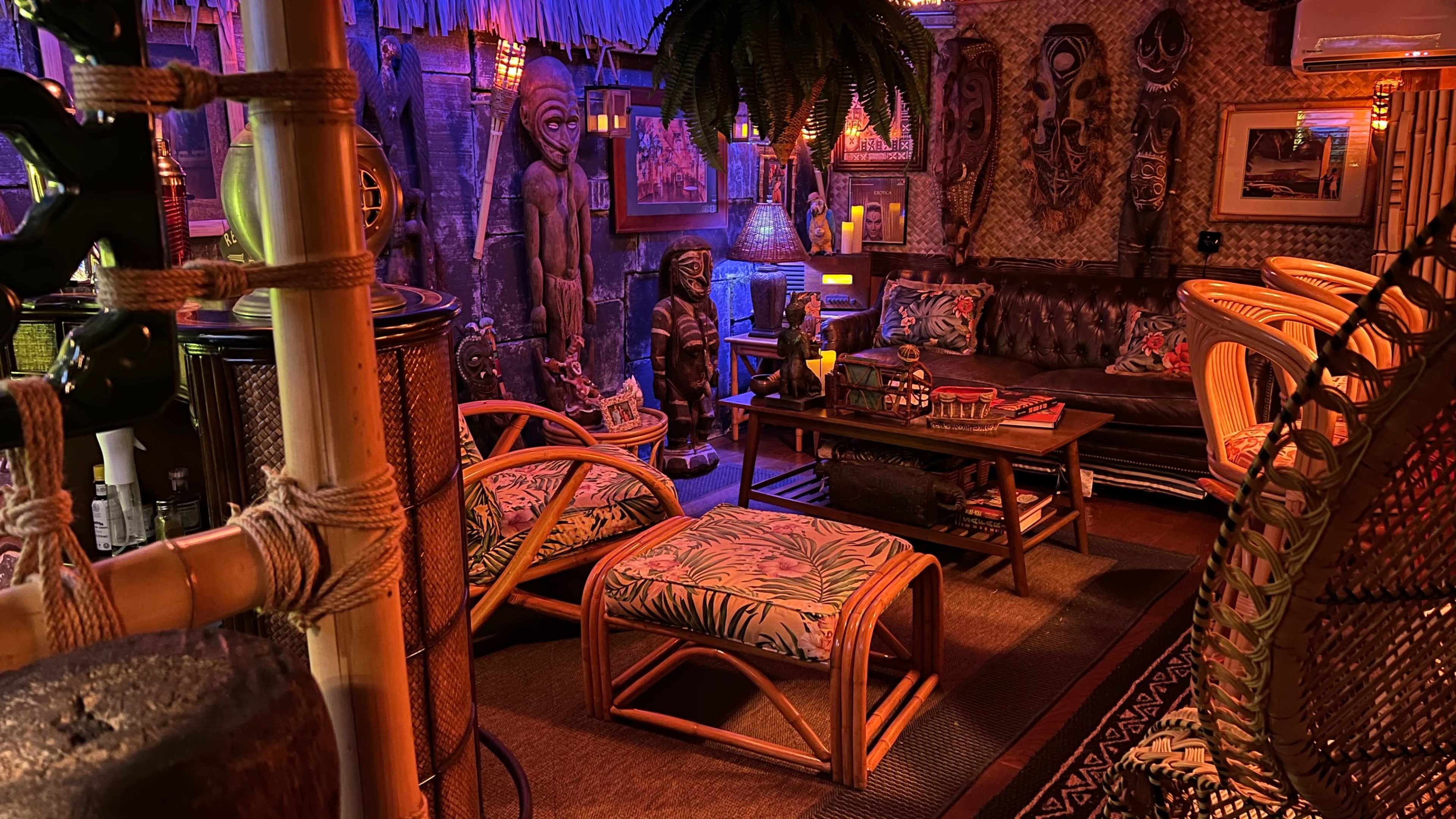 The image displays a tiki-themed interior with wooden sculptures, patterned furniture, and dim lighting creating a tropical atmosphere.