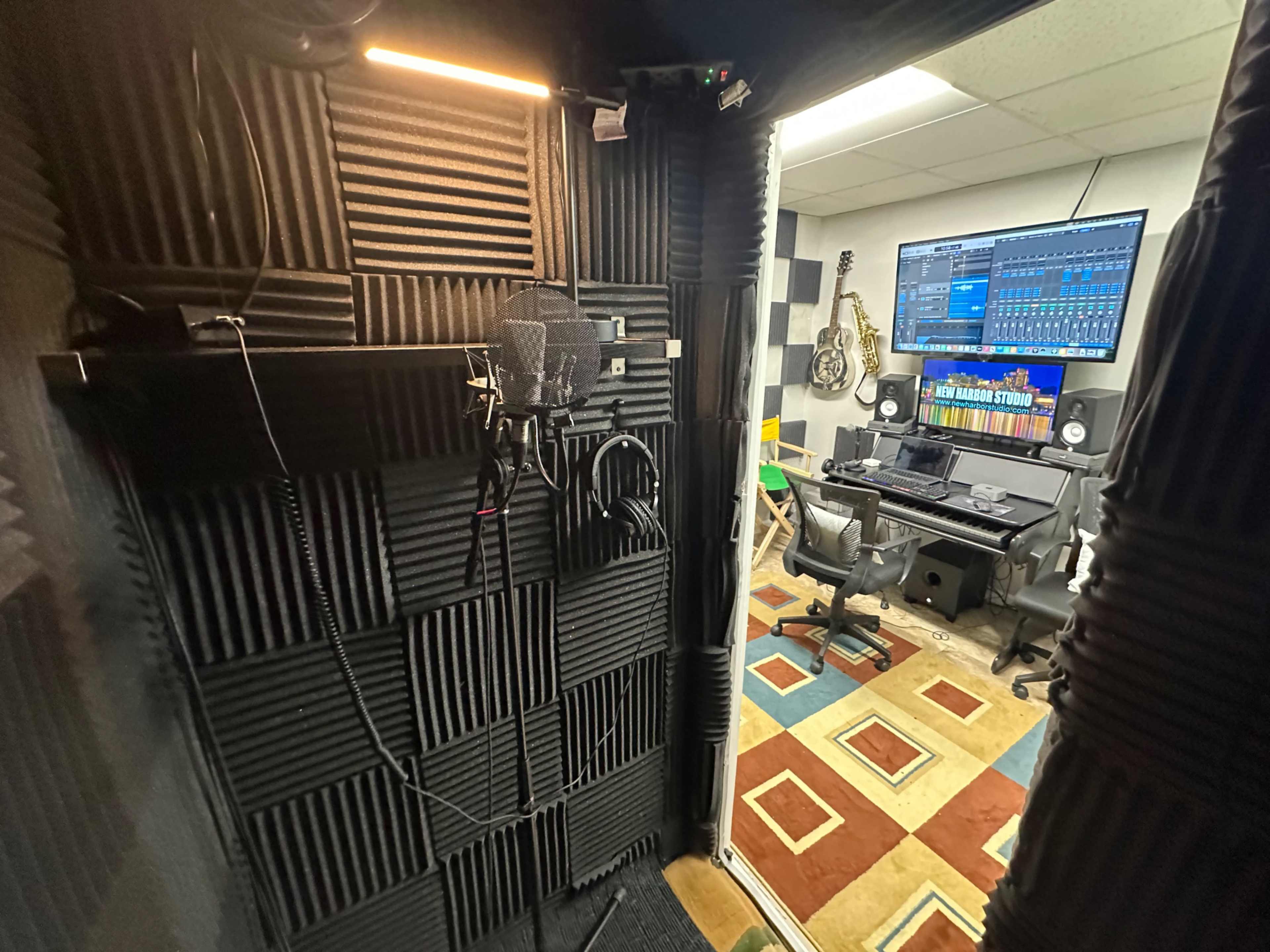 The image shows a soundproof recording booth with a microphone setup on one side and a control area featuring a computer and musical instruments visible in the background.