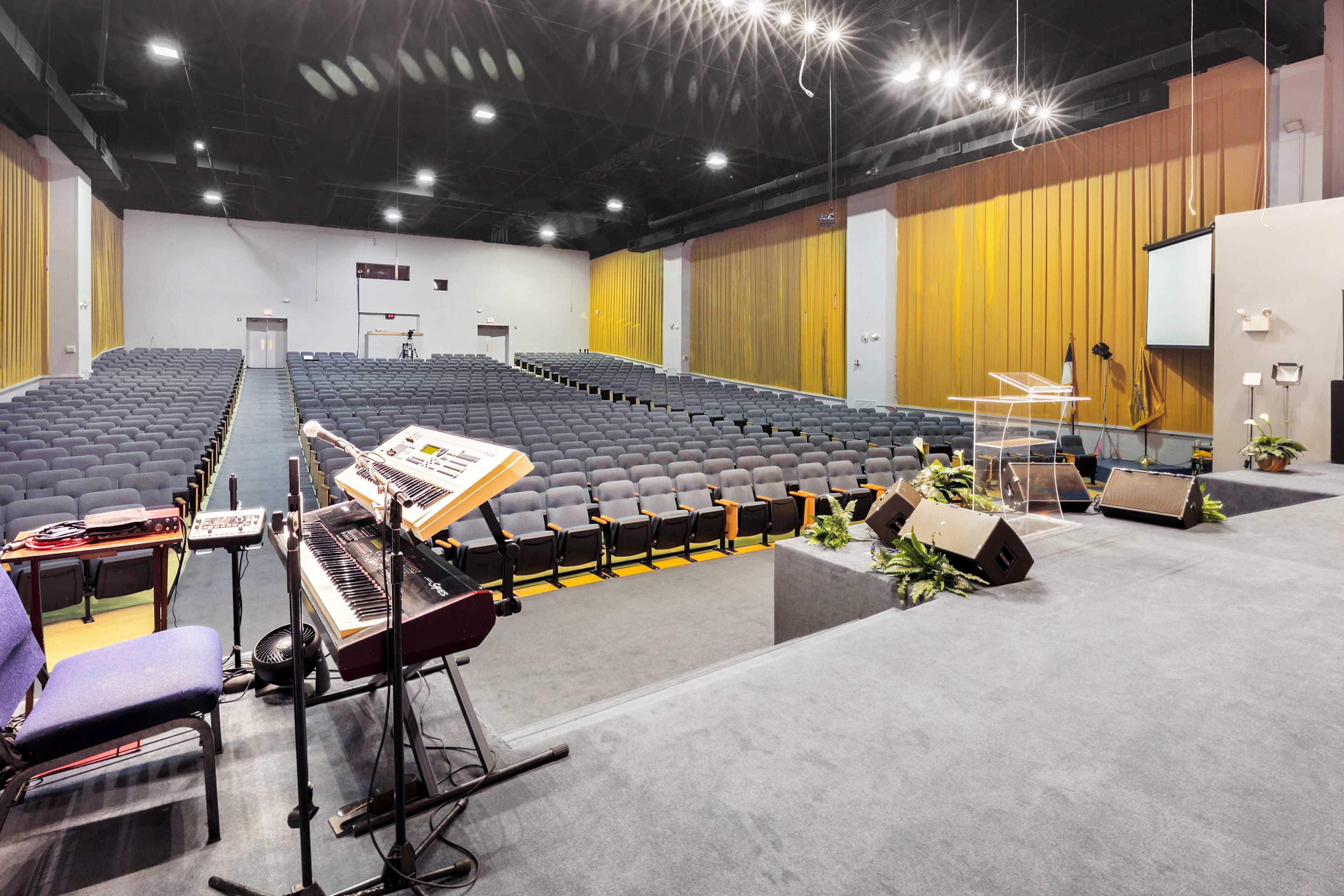 A spacious auditorium with rows of empty chairs, a keyboard setup, and a podium on a stage.