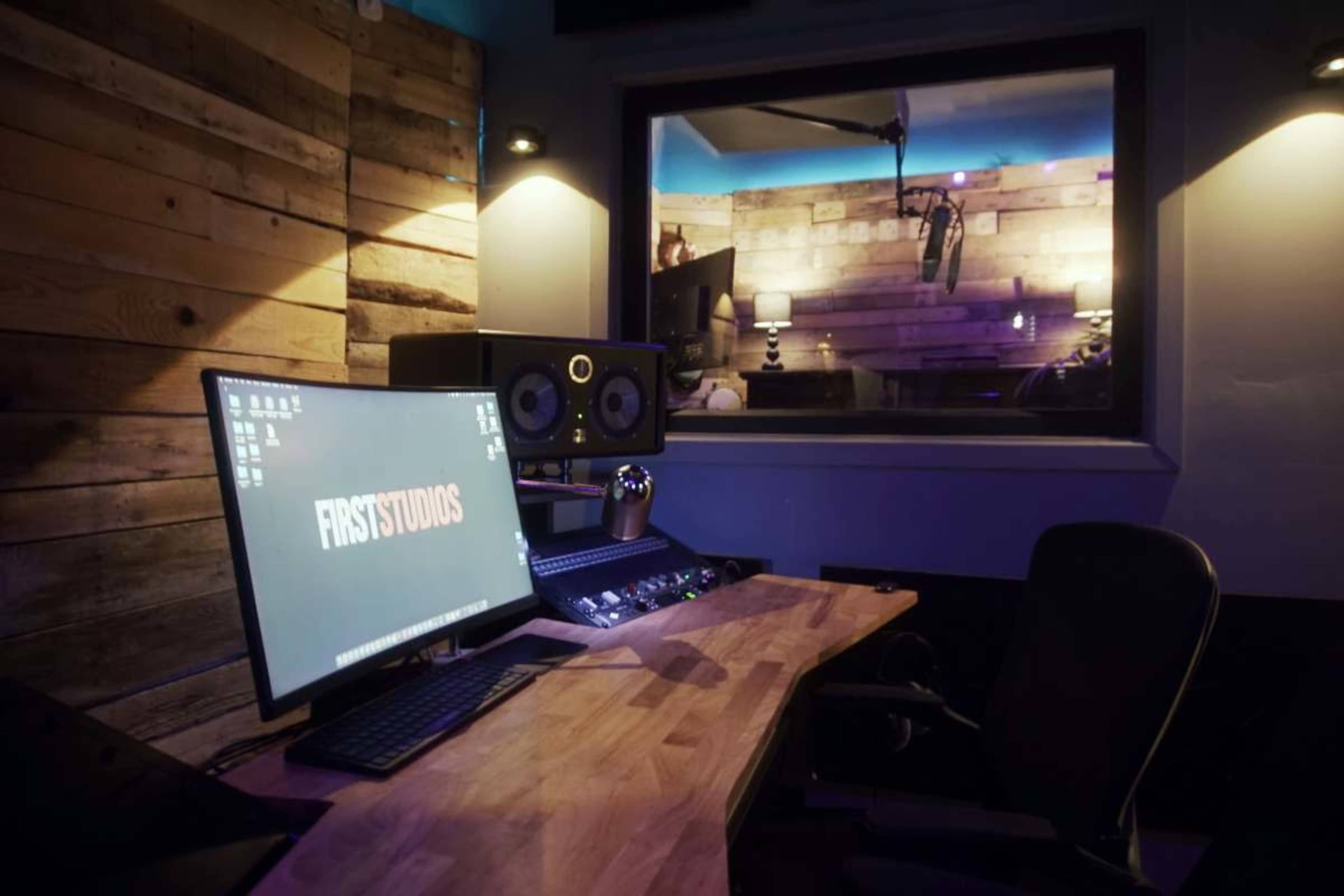 The image shows a recording studio with a wooden desk, computer, and audio equipment, featuring a glass-walled control room in the background.