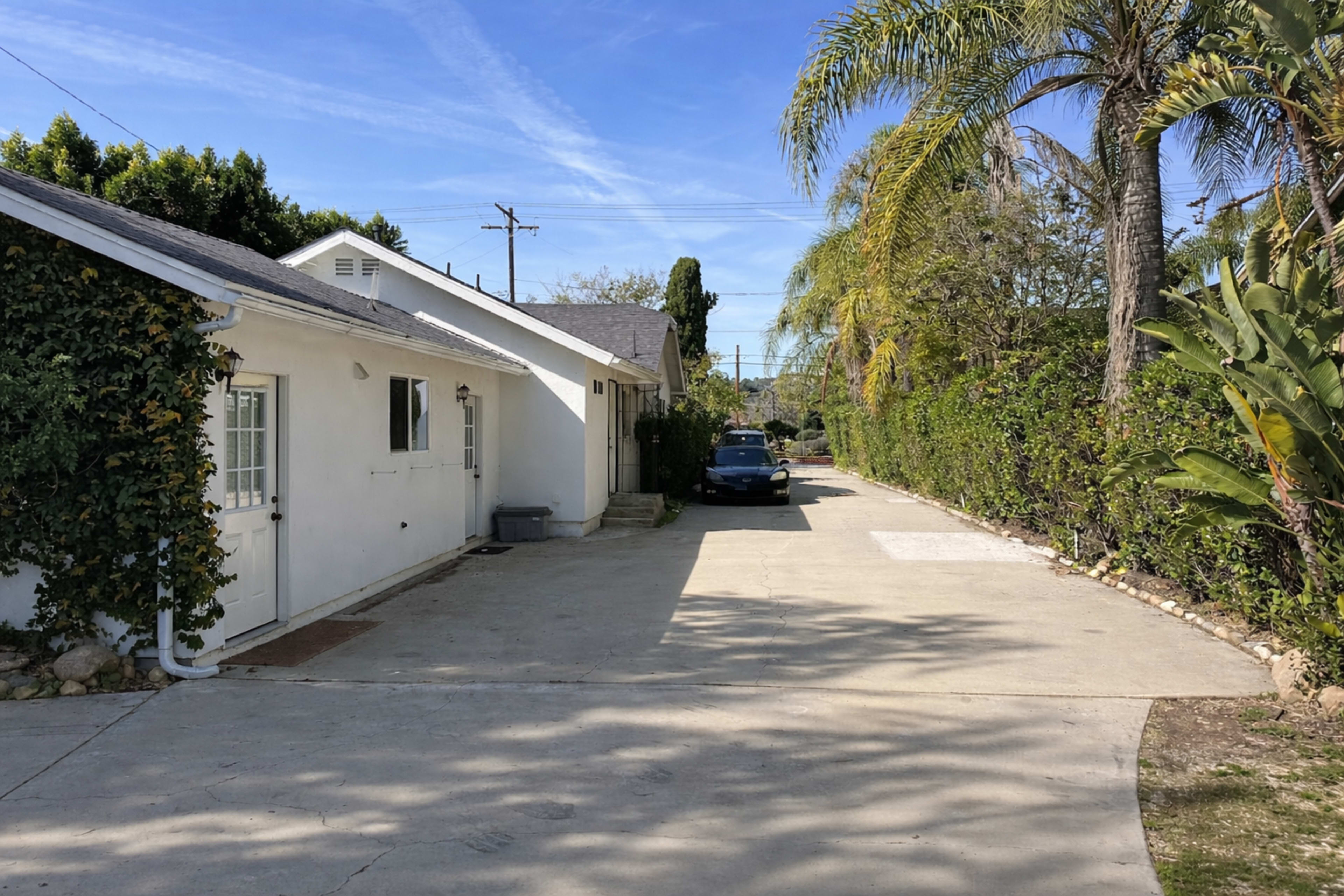 The image shows a concrete driveway leading between two greenery-covered structures, with a blue car parked to the right under clear blue skies.