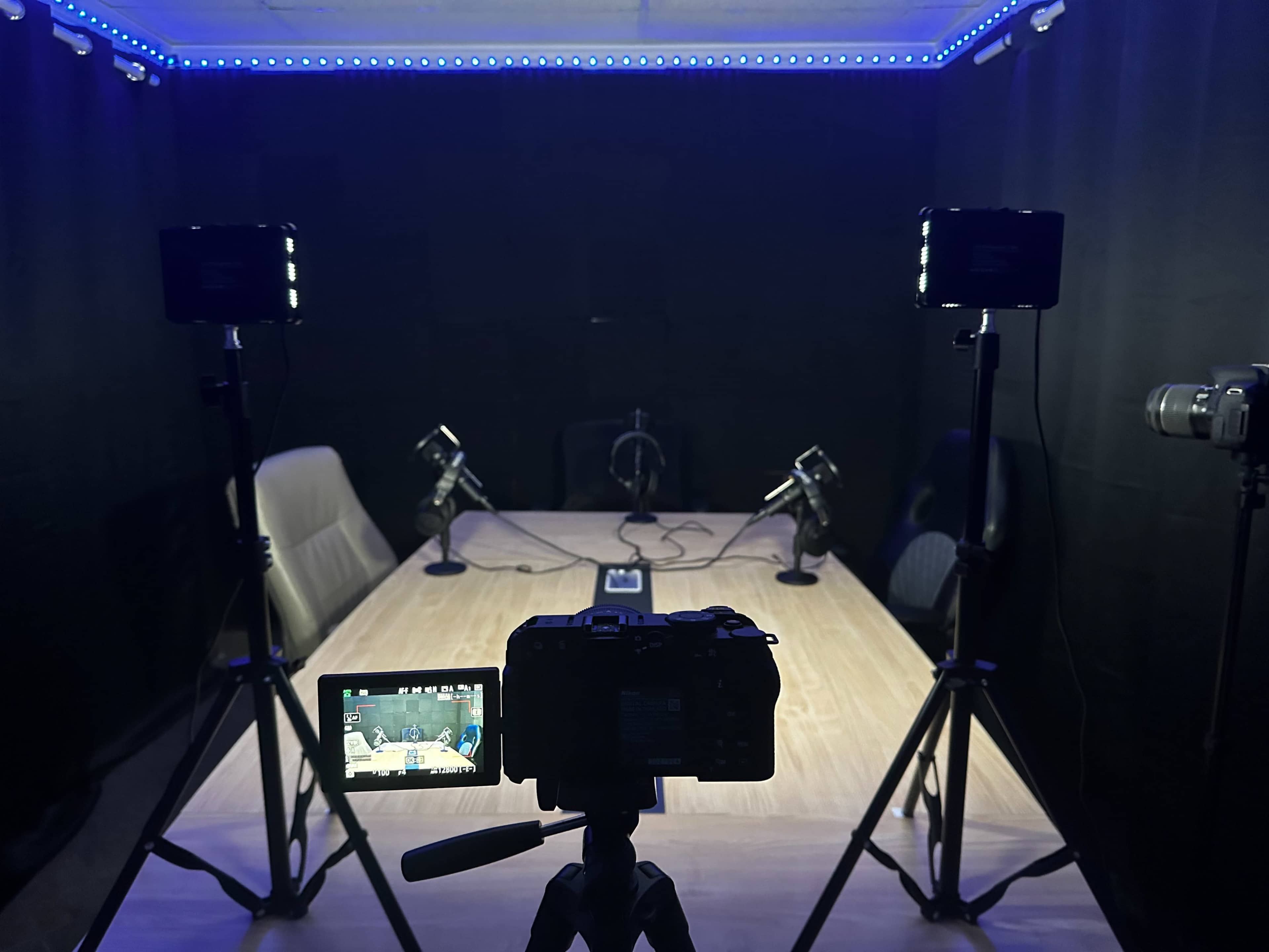 A camera and lights are set up in a dark studio featuring a large wooden table surrounded by chairs and microphones.