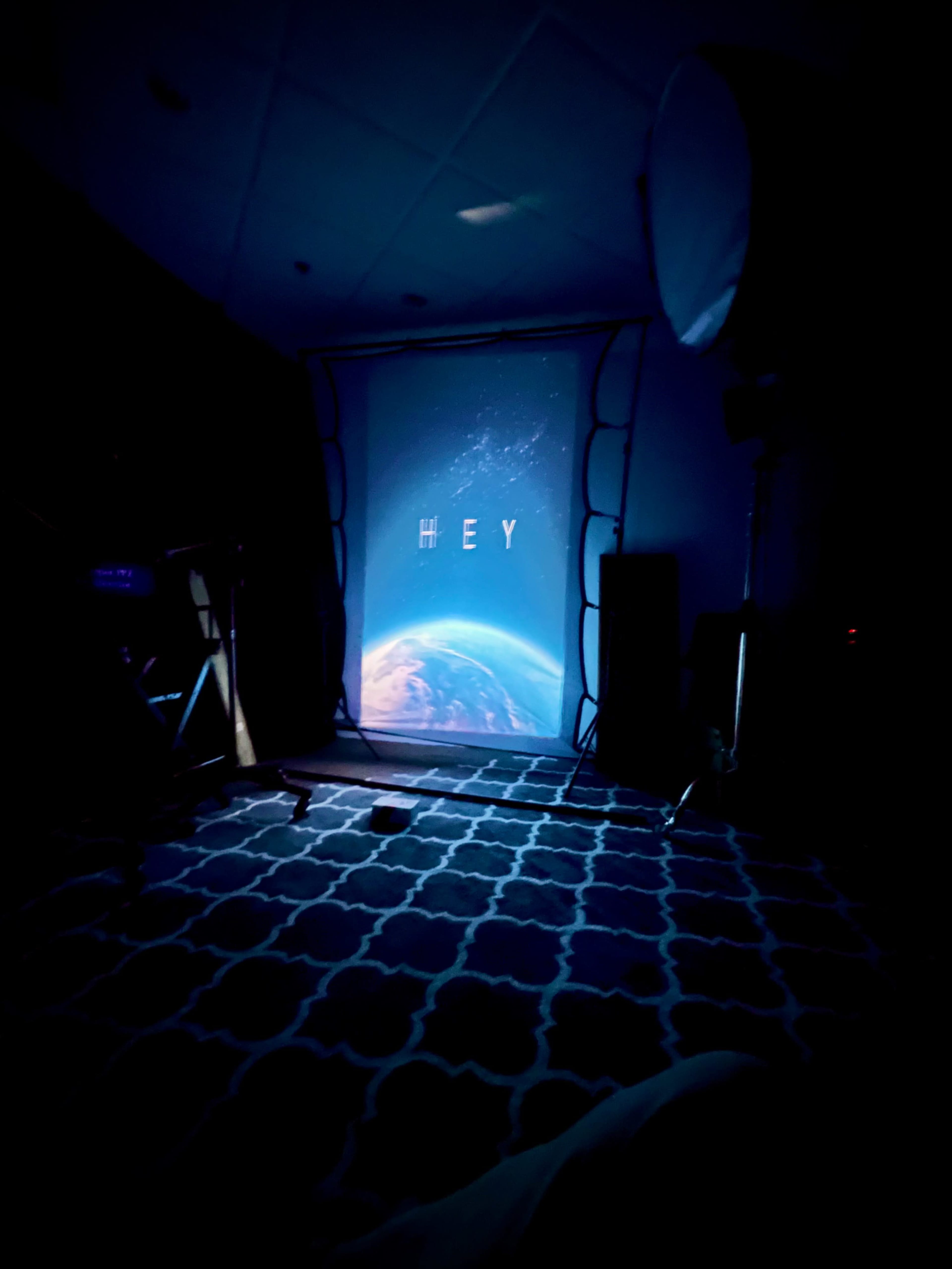 A dimly lit room with a projector displaying the word "HEY" over an image of Earth.