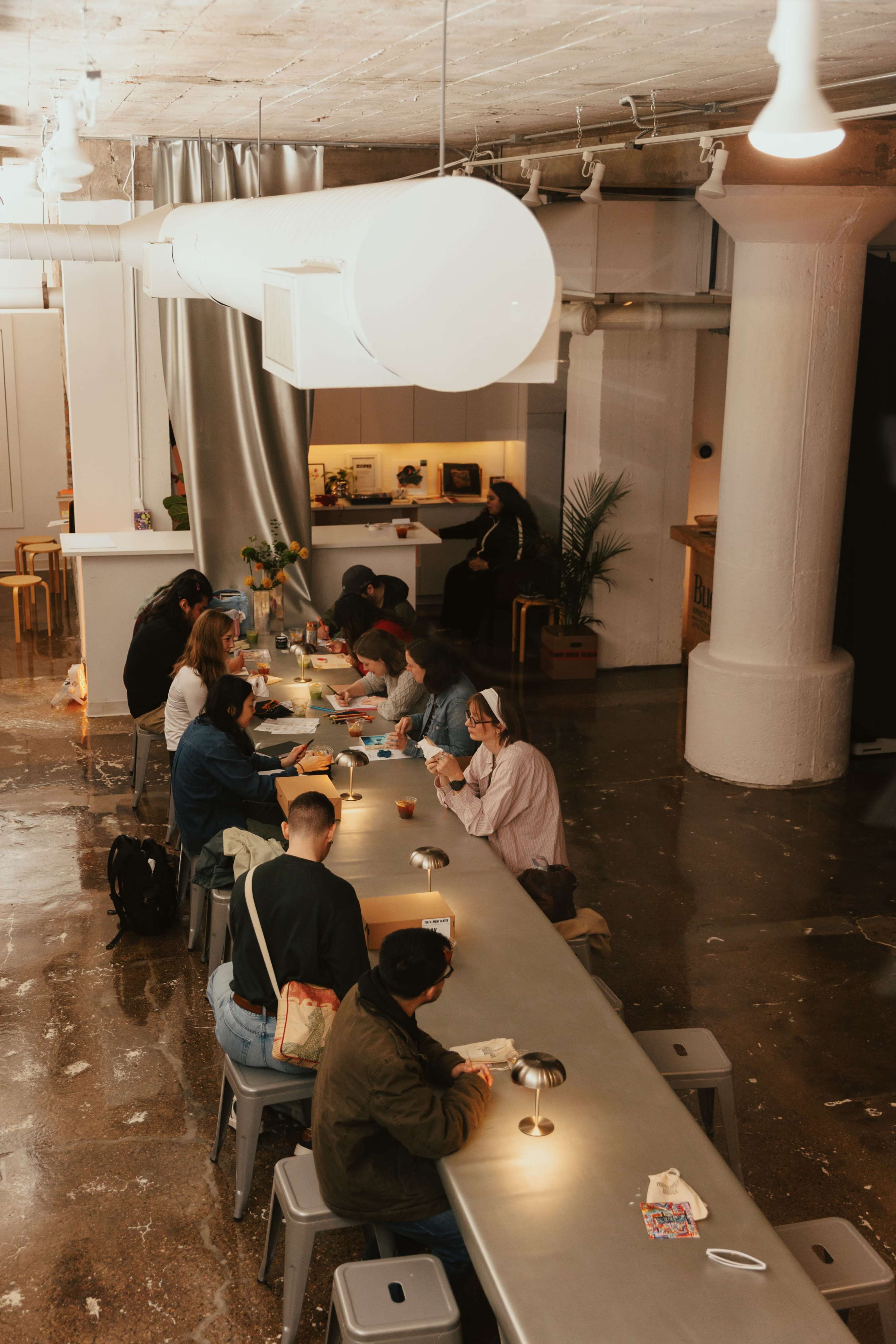A group of people is seated at a long table in a modern cafe, engaged in conversations while enjoying drinks and food.