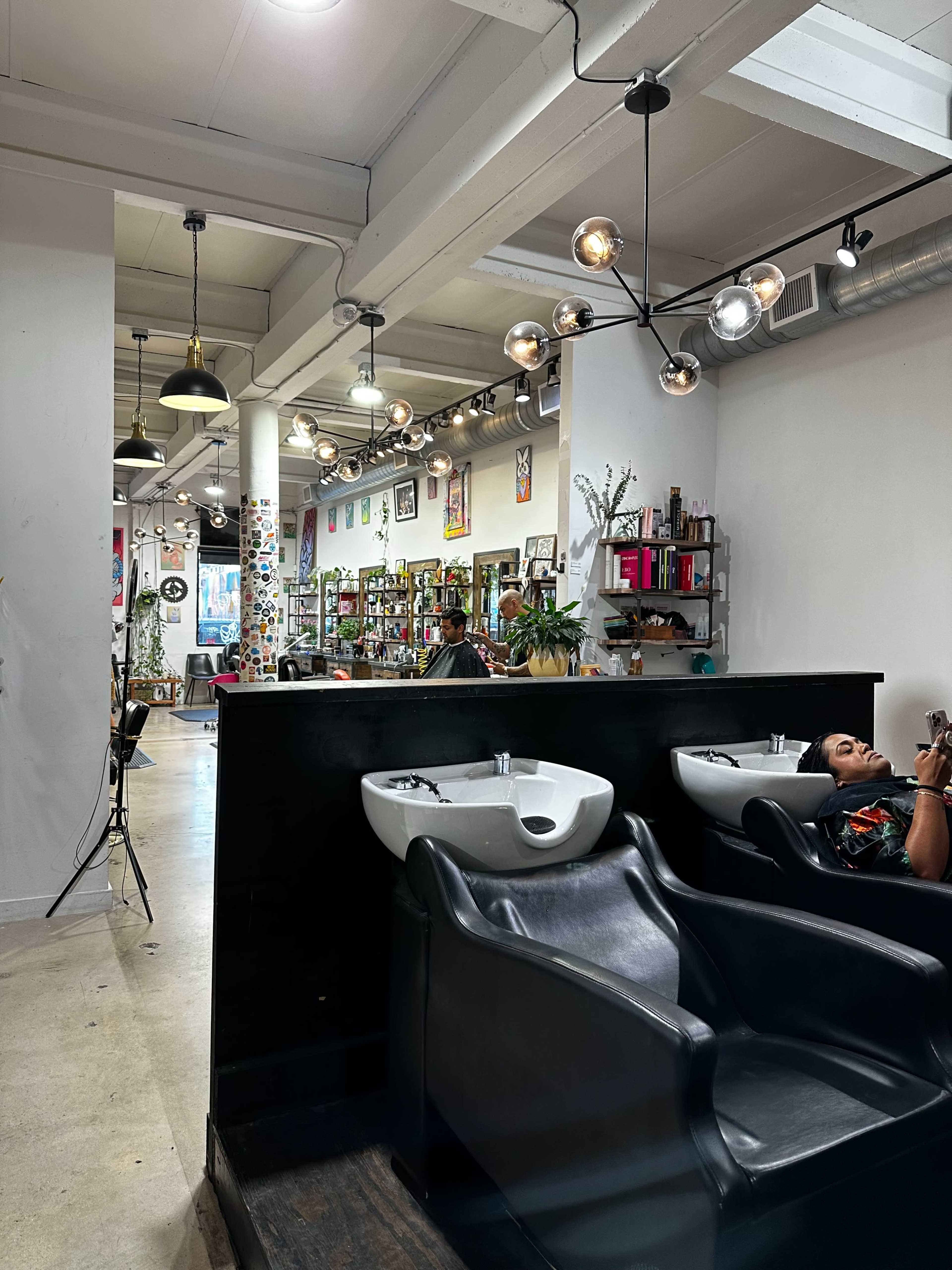 The image shows the interior of a modern hair salon with black styling chairs, washing stations, and shelves lined with hair products.