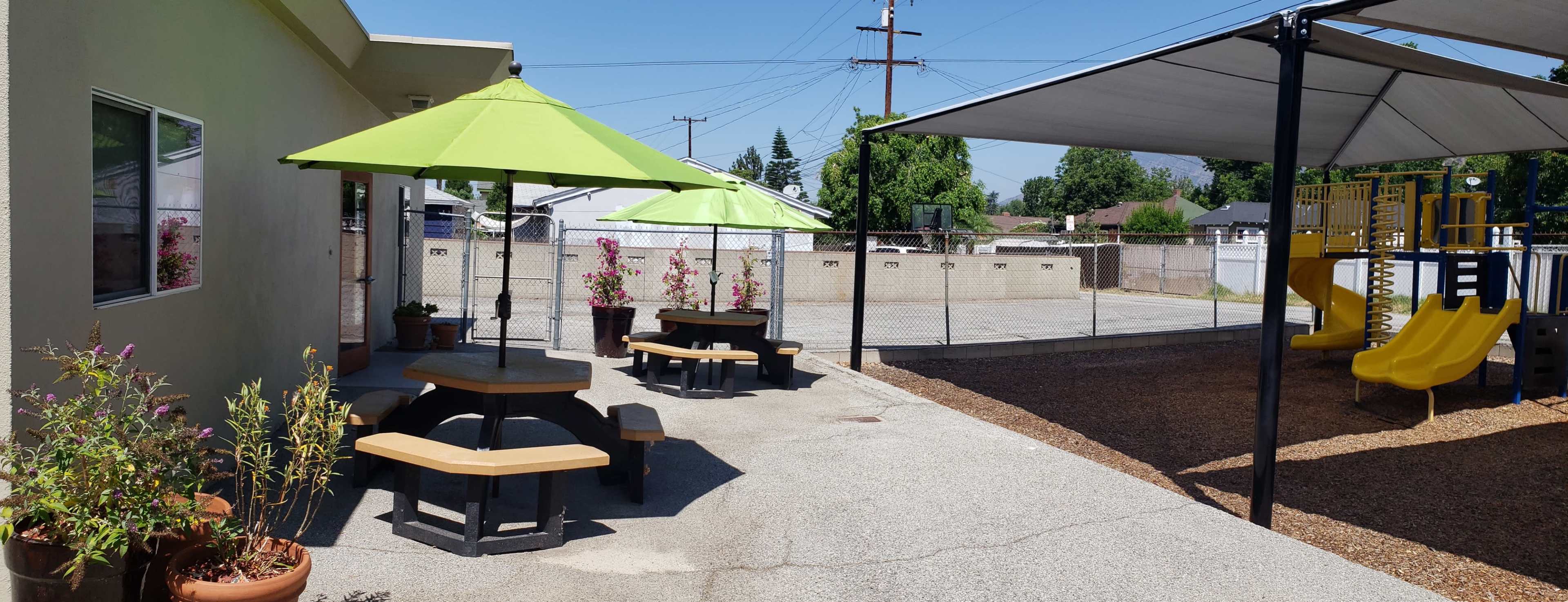 The image shows a playground area with yellow slides and two green umbrellas over picnic tables, surrounded by a fence and featuring flower pots nearby.