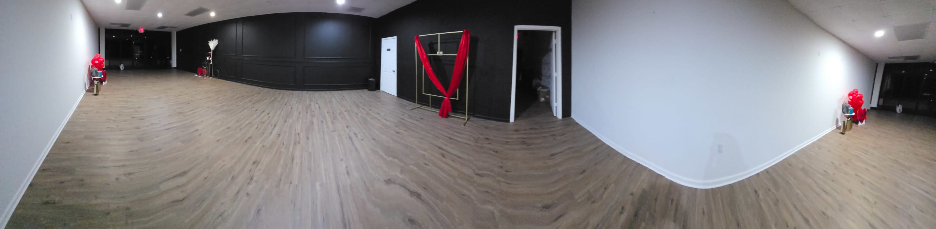 The image shows a spacious, empty room with a polished wooden floor, adorned with red decorations on one side and a door leading to another area on the right.