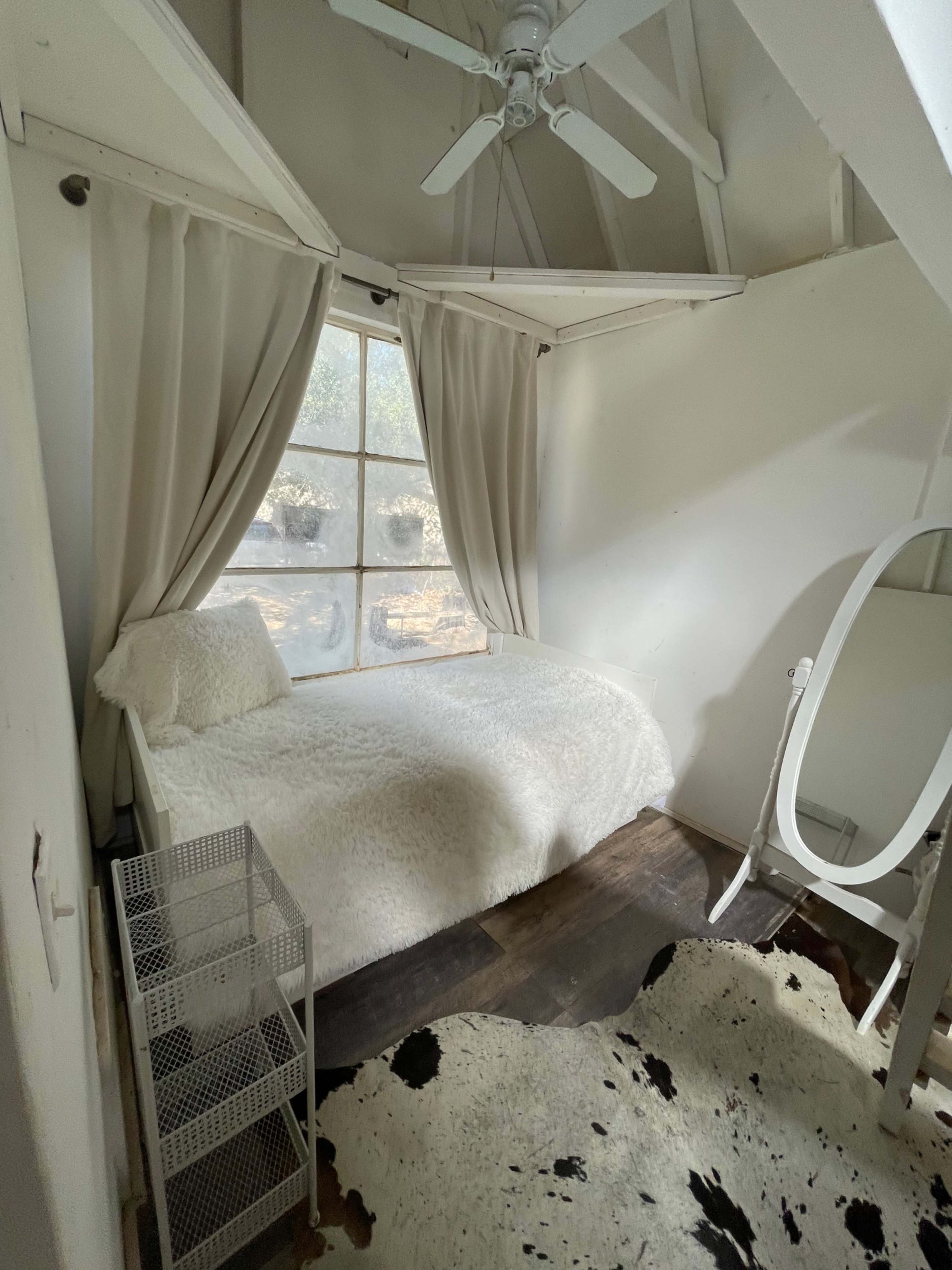 A small bedroom with a single bed, a white mirror, a wire storage unit, and a window with light curtains, set on wooden flooring with a cowhide rug.