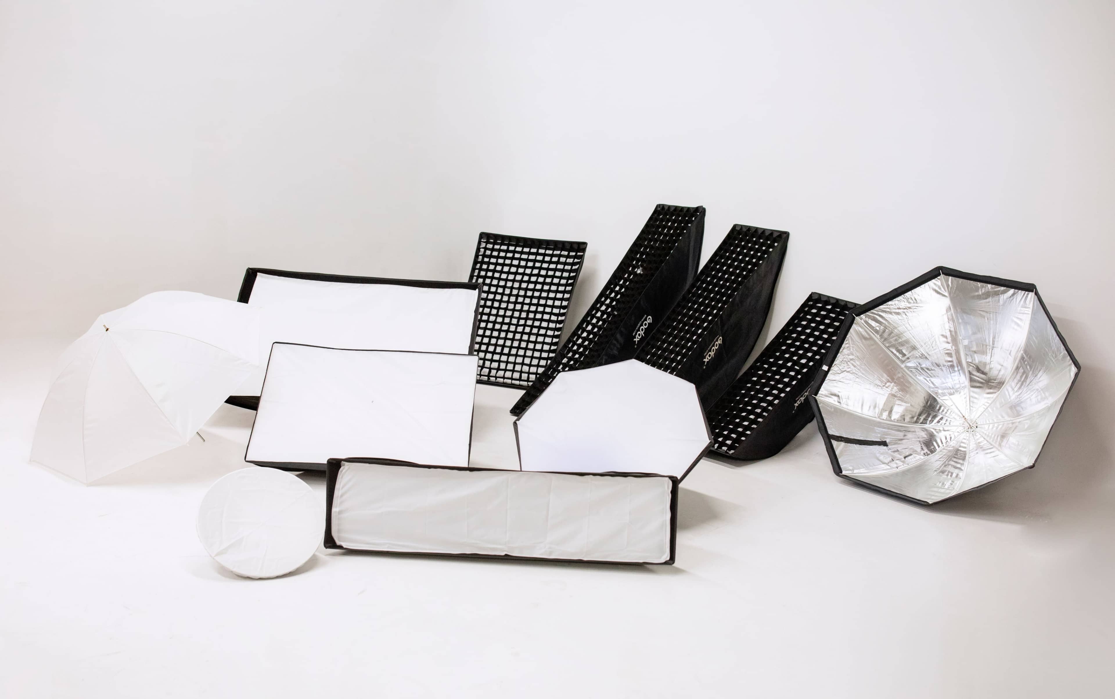 The image displays various photography lighting modifiers arranged on a neutral background.