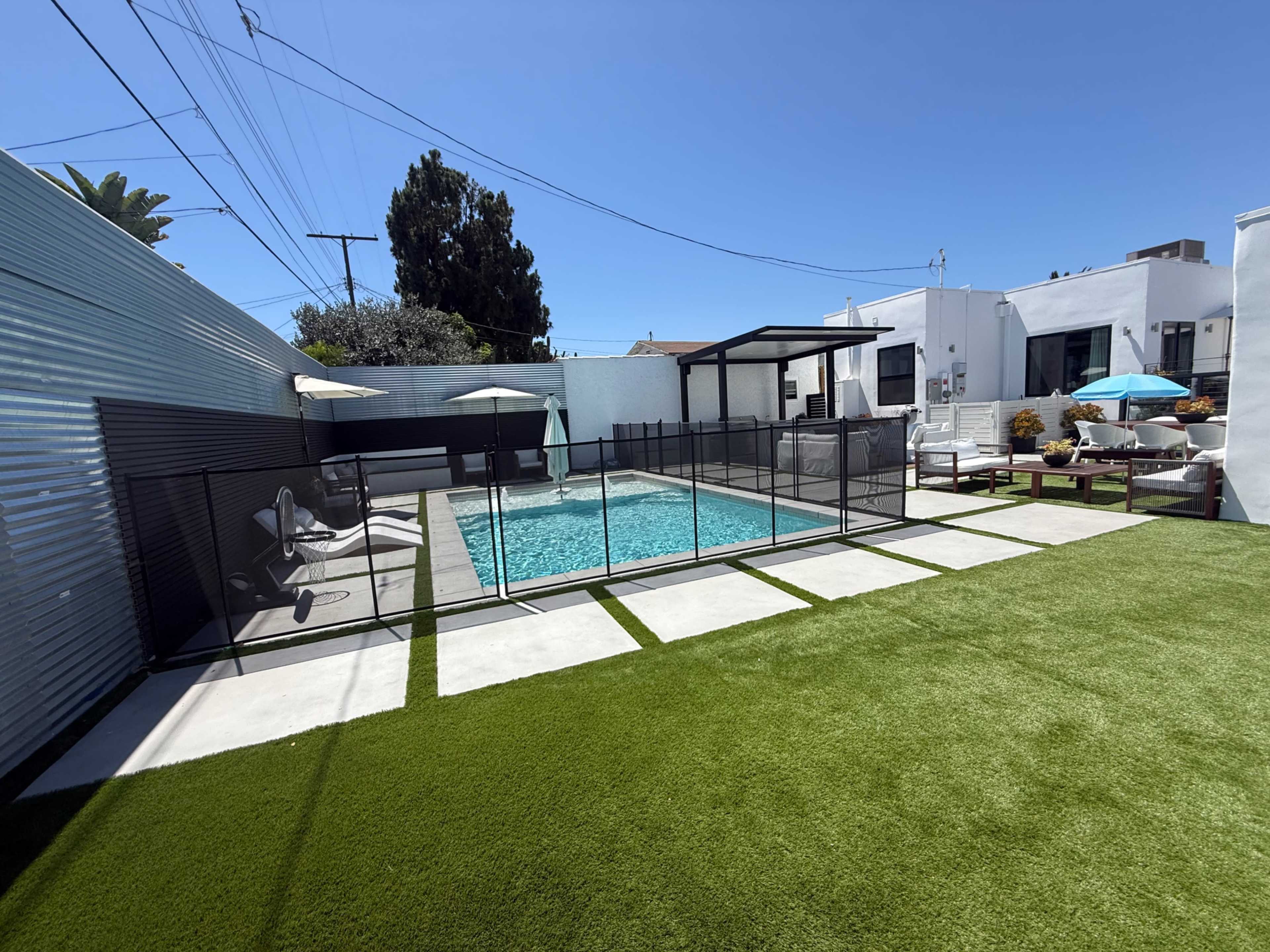 The image shows a backyard pool area featuring a fenced-in swimming pool surrounded by artificial grass and lounge furniture.