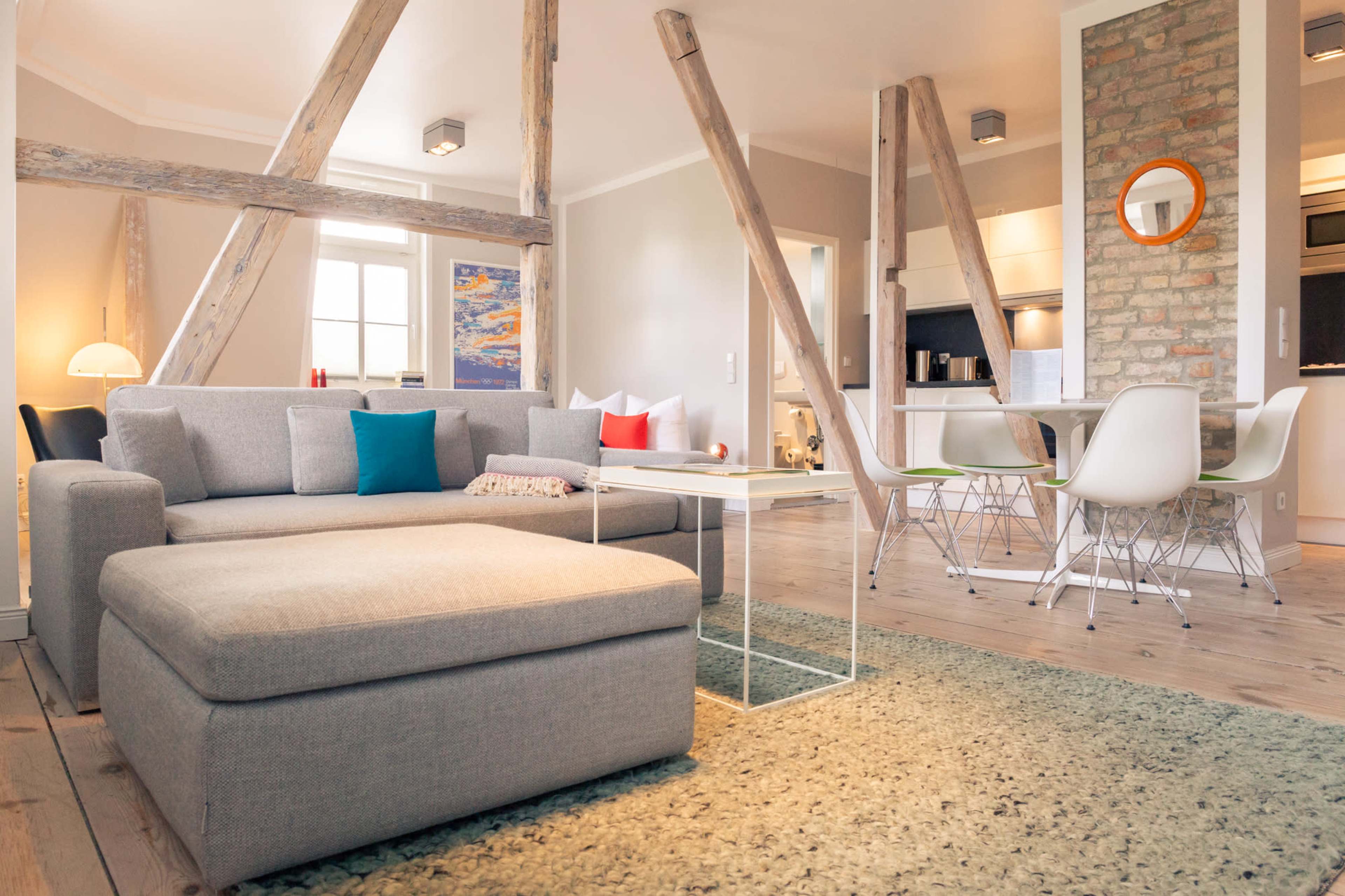 A modern living room with a gray sectional sofa, a coffee table, and an open kitchen area, featuring wooden beams and a textured rug.