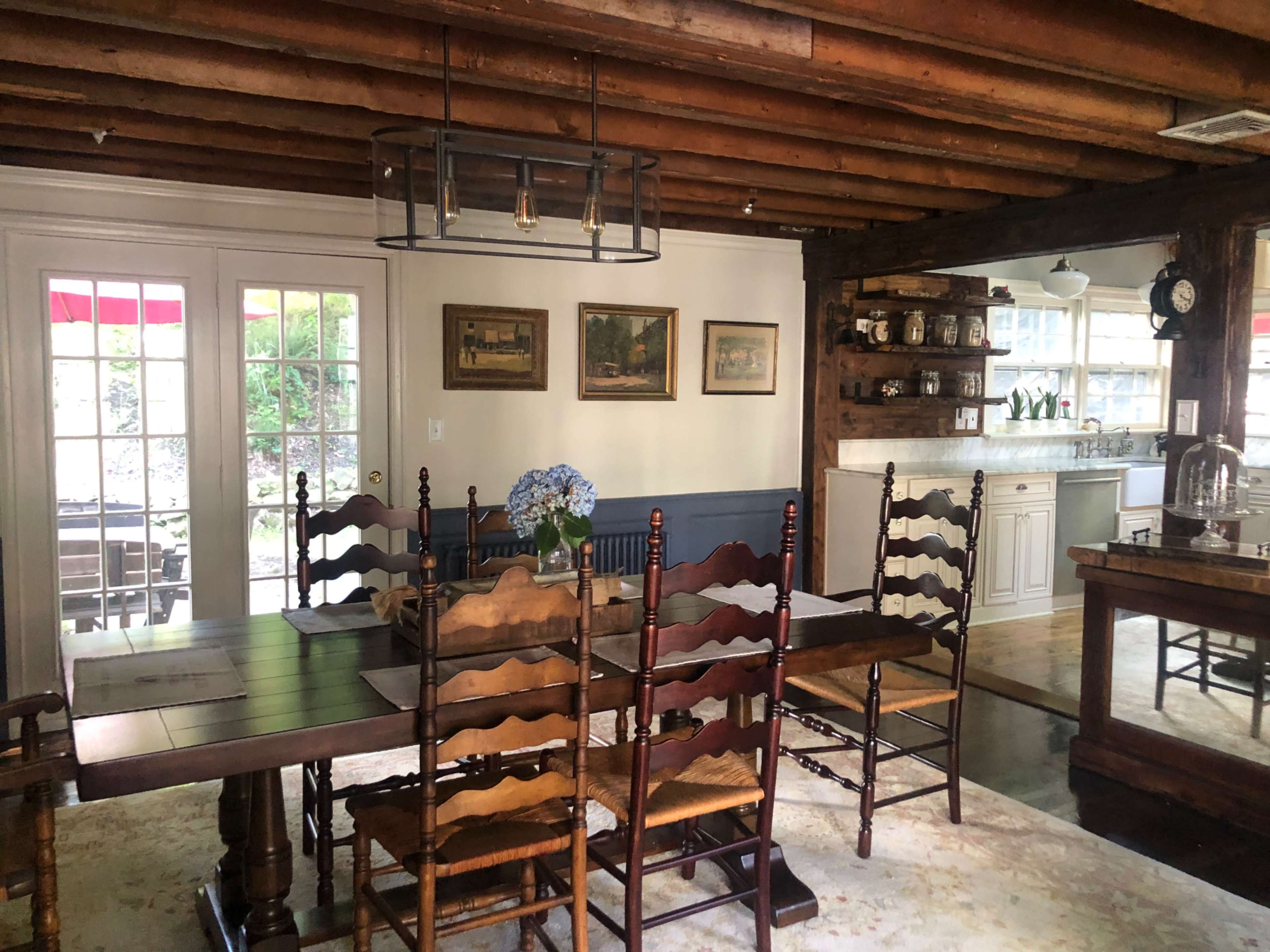 A dining area with a wooden table surrounded by chairs, featuring a floral centerpiece and visible wood beam ceiling, adjoining a kitchen with cabinets and a counter.