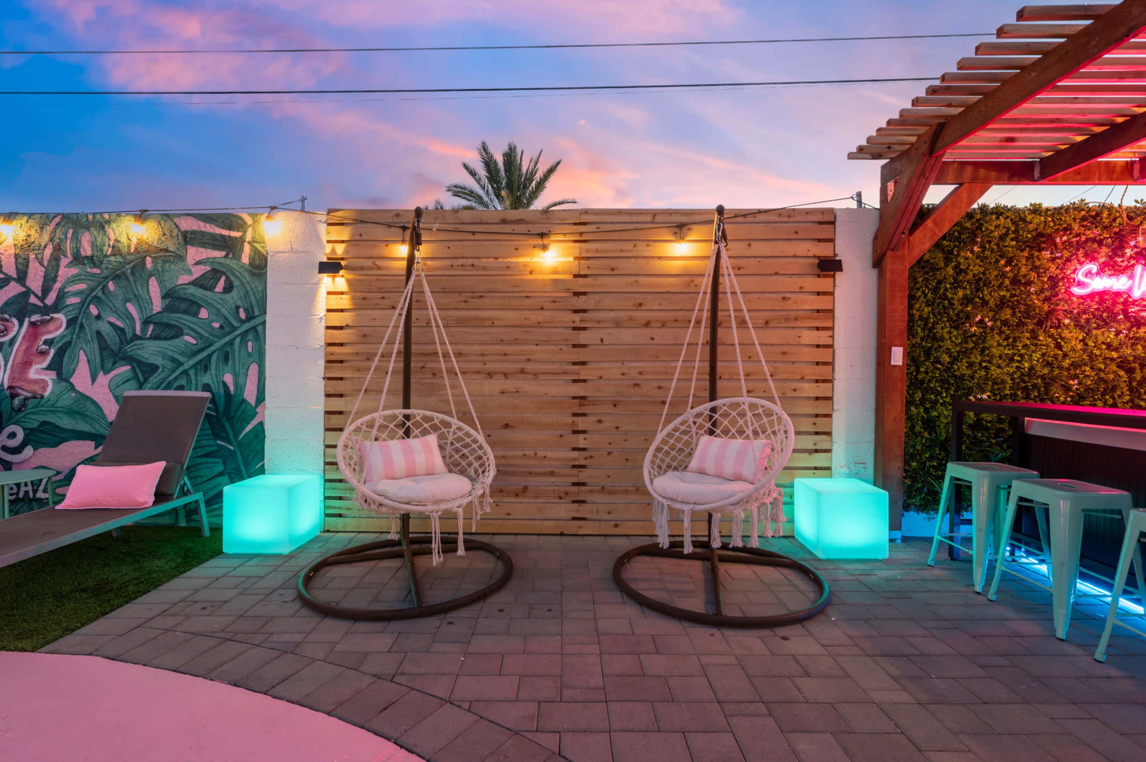 The image features a modern outdoor space with two hanging chairs, illuminated by colored lights, against a backdrop of a wooden wall and greenery.