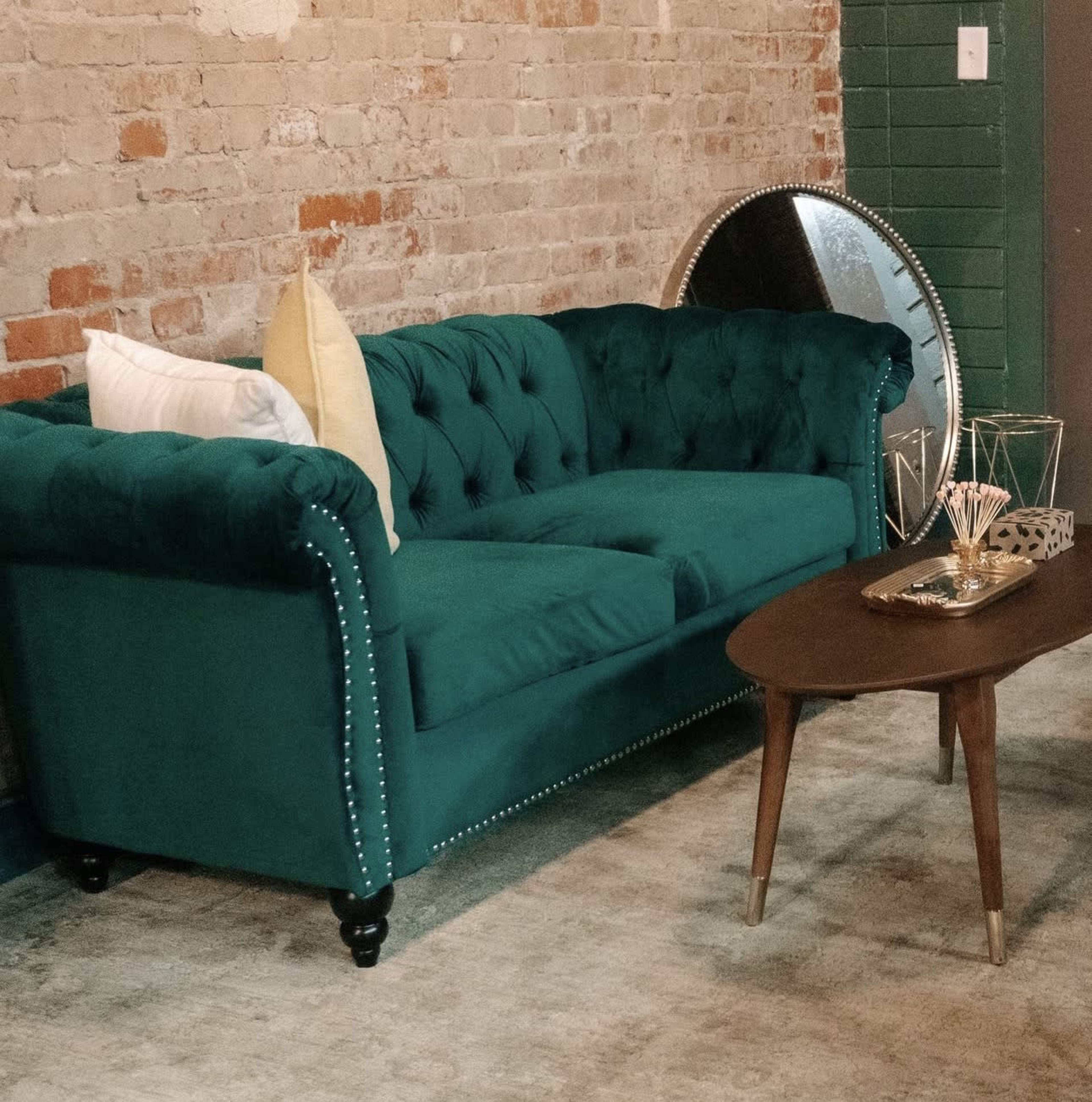 The image shows a teal tufted couch with decorative pillows, accompanied by a wooden table and a round mirror against a textured brick wall.
