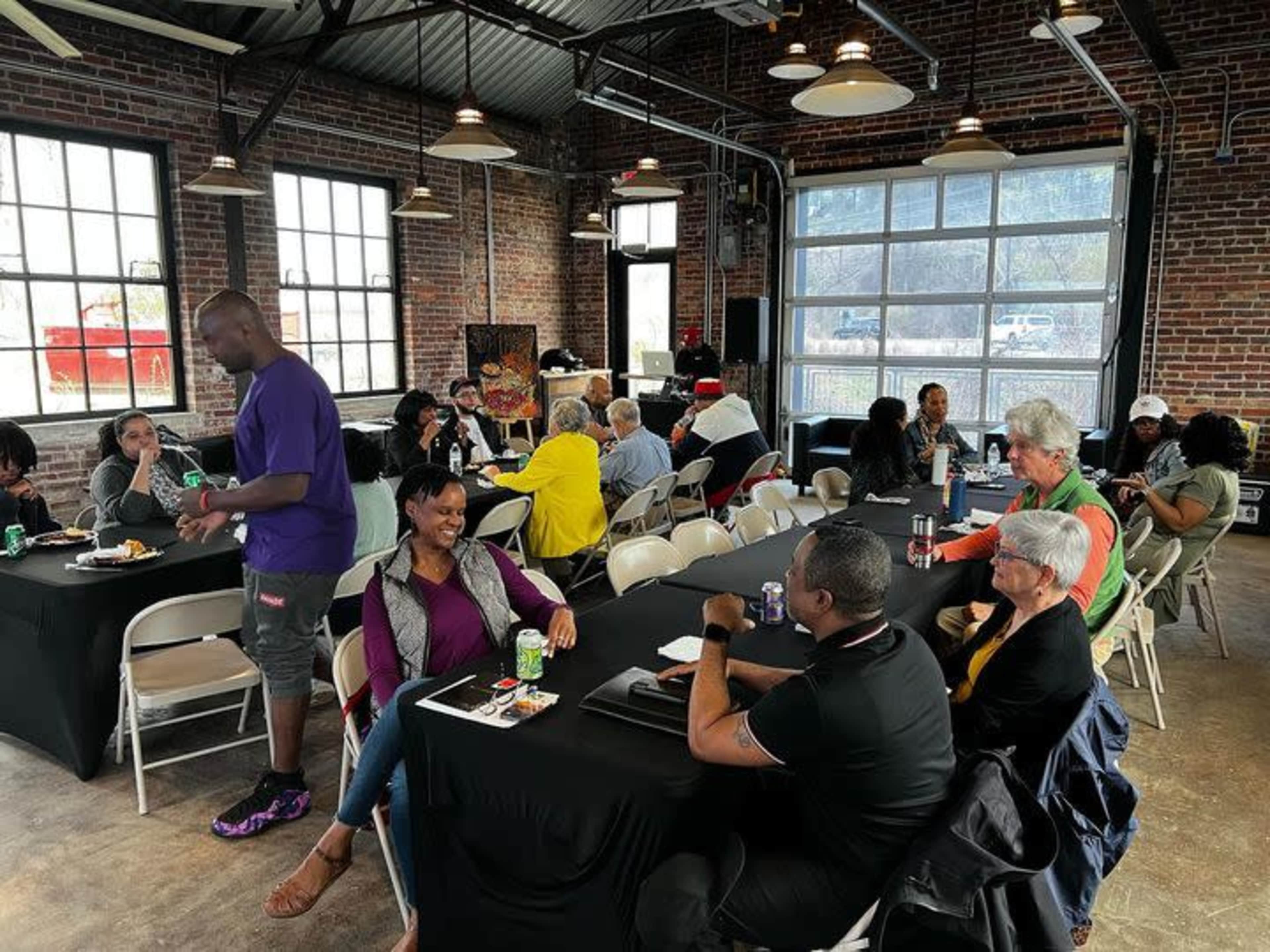 A group of people sits at tables in a spacious industrial-style venue with large windows and exposed brick walls.