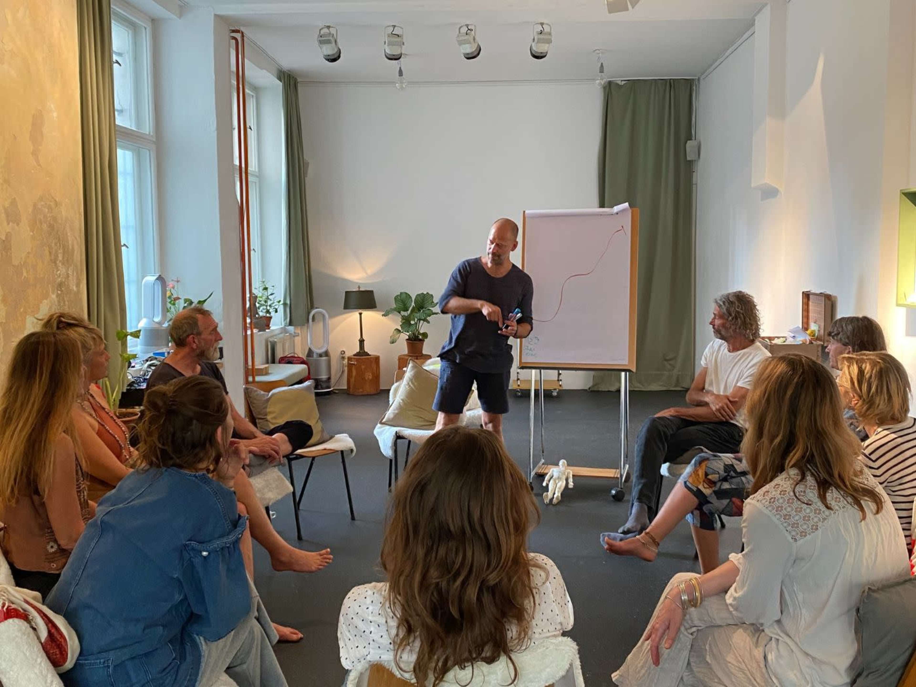 A group of people sits in a circle in a well-lit room, with one person standing and presenting to the group using a flip chart.