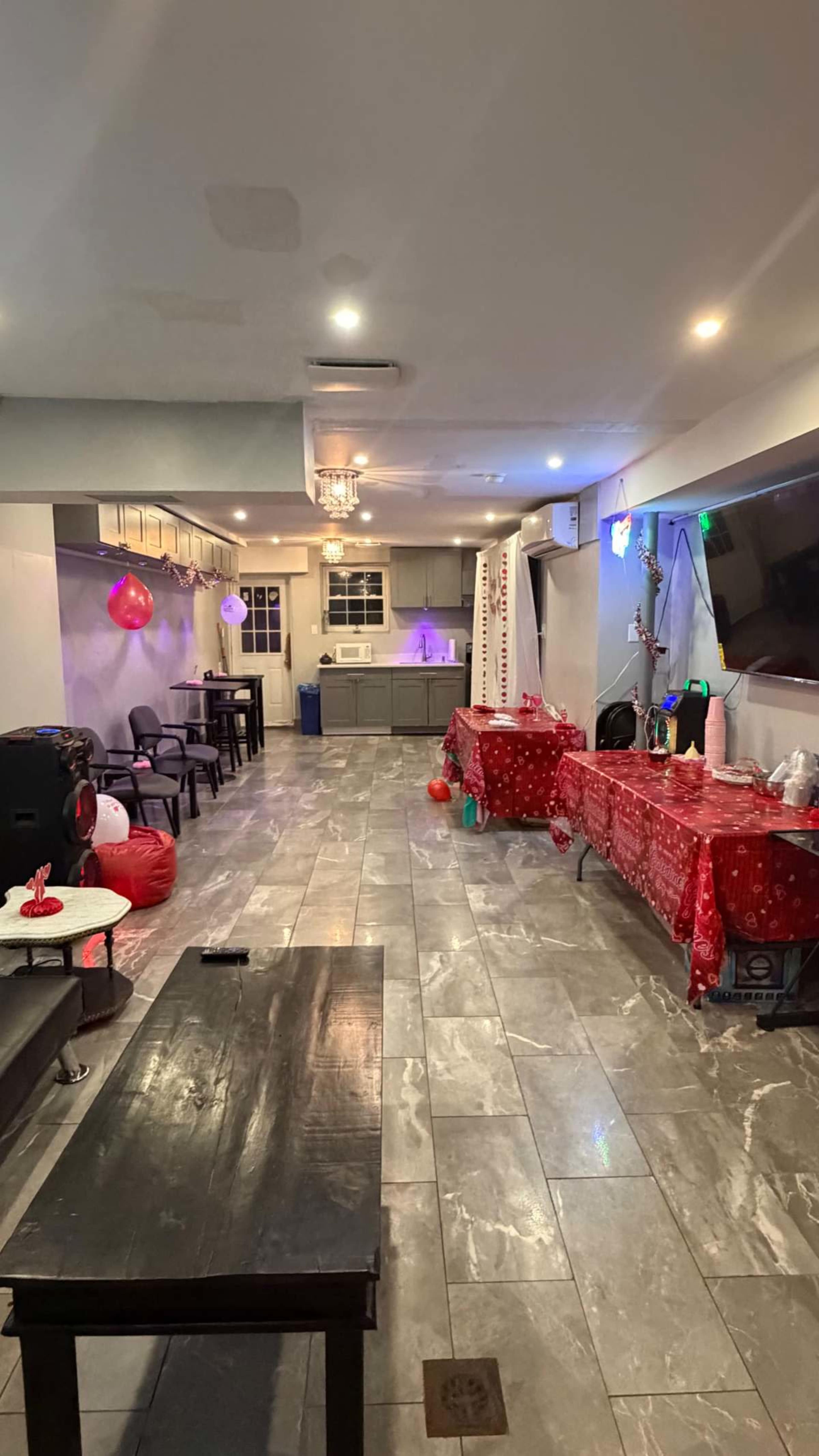 The image shows a decorated indoor space with tables covered in red tablecloths, chairs, and a kitchen area in the background, all set up for a celebration.