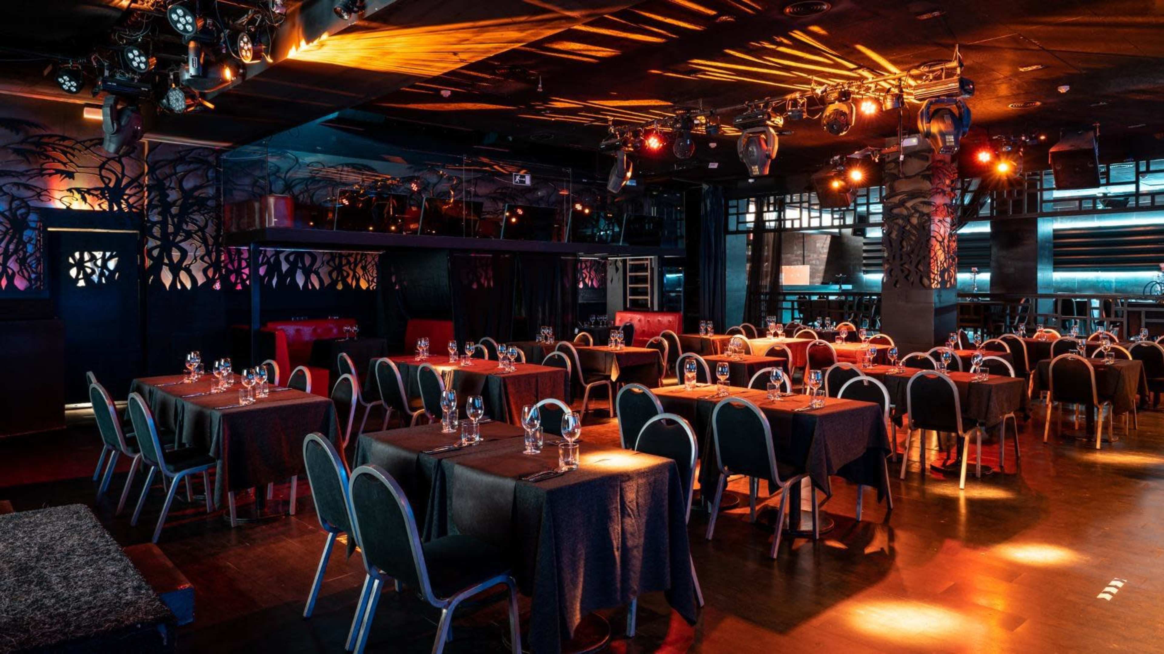 The image shows a dimly lit restaurant with tables set for dining, featuring a stage area and various overhead lights.