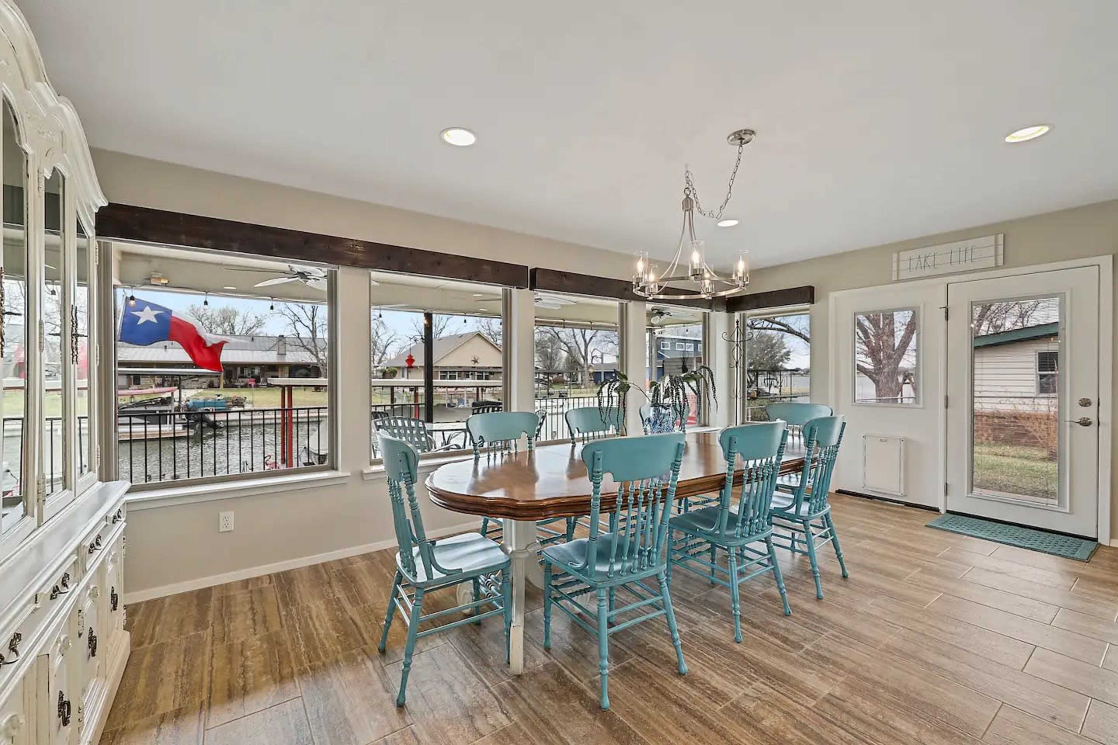 A dining area with a wooden table surrounded by blue chairs, large windows, and a door leading outside, featuring a Texas flag visible through the window.