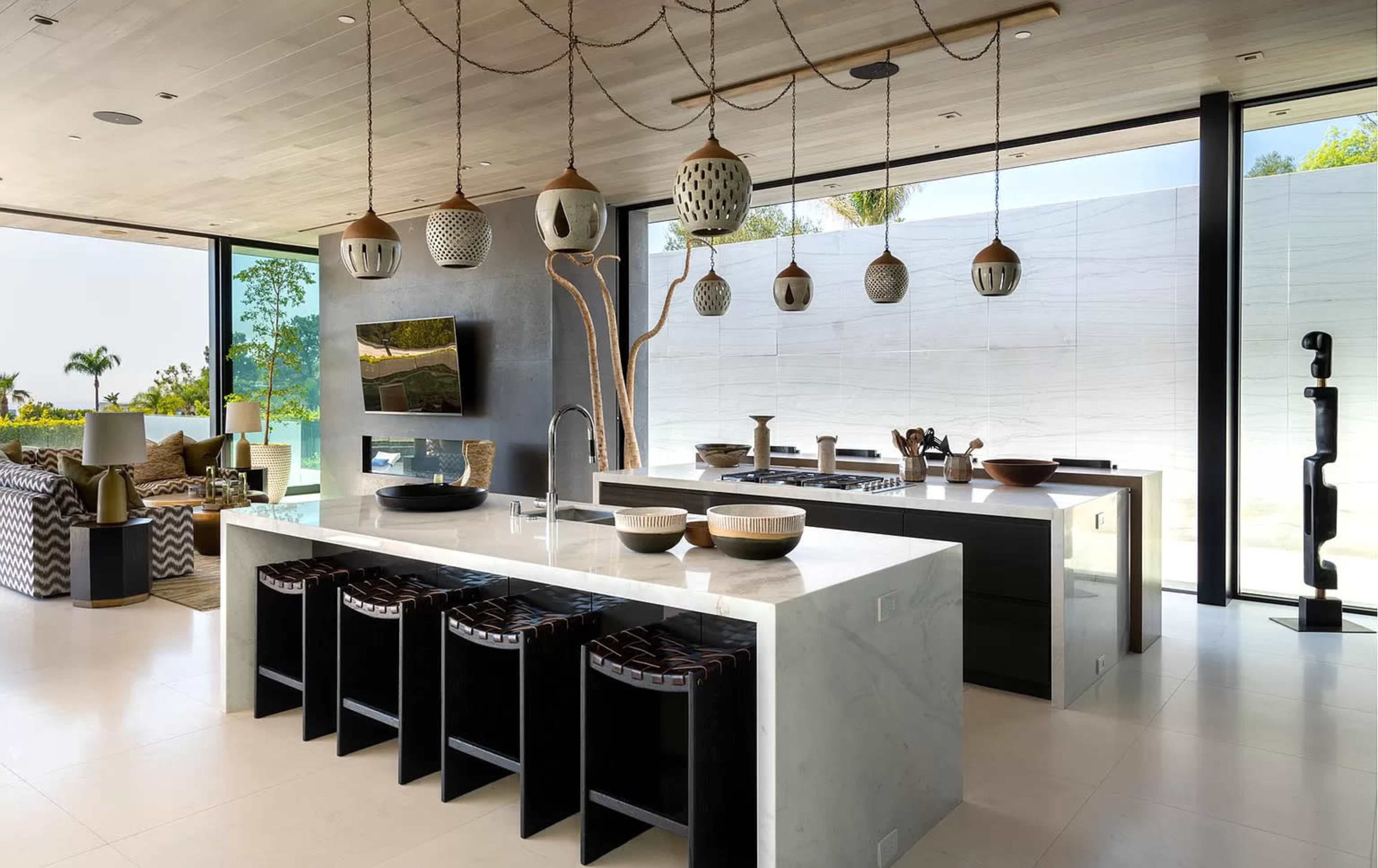 The image shows a modern kitchen featuring a large white stone island with dark seating, stylish pendant lights, and a backdrop of glass walls revealing an outdoor view.