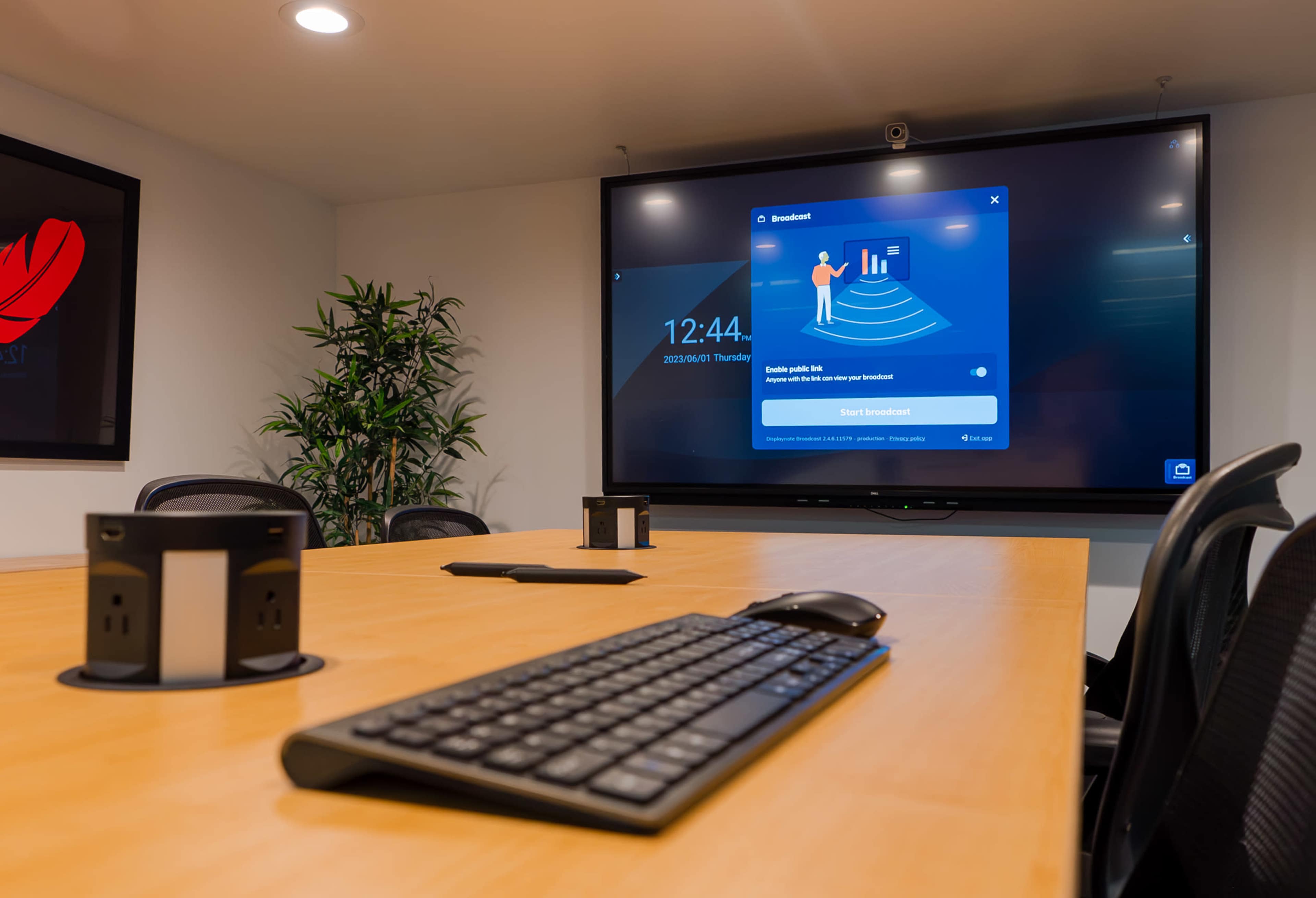 A modern conference room features a large screen displaying a broadcast interface, with a wooden table and a keyboard in the foreground.