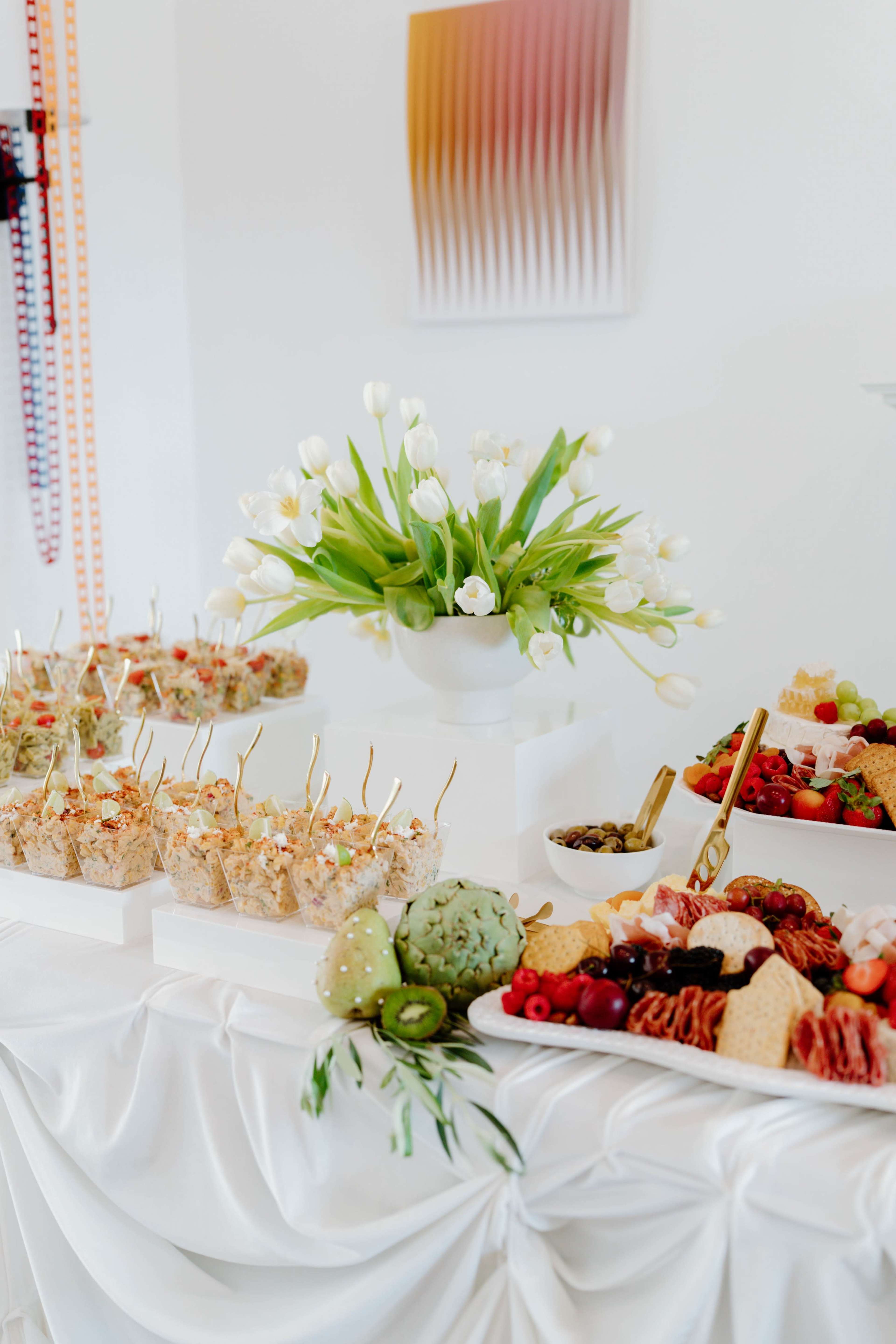 A table is arranged with an assortment of elegantly presented food platters, including appetizers and fresh fruits, surrounded by a centerpiece of white tulips.