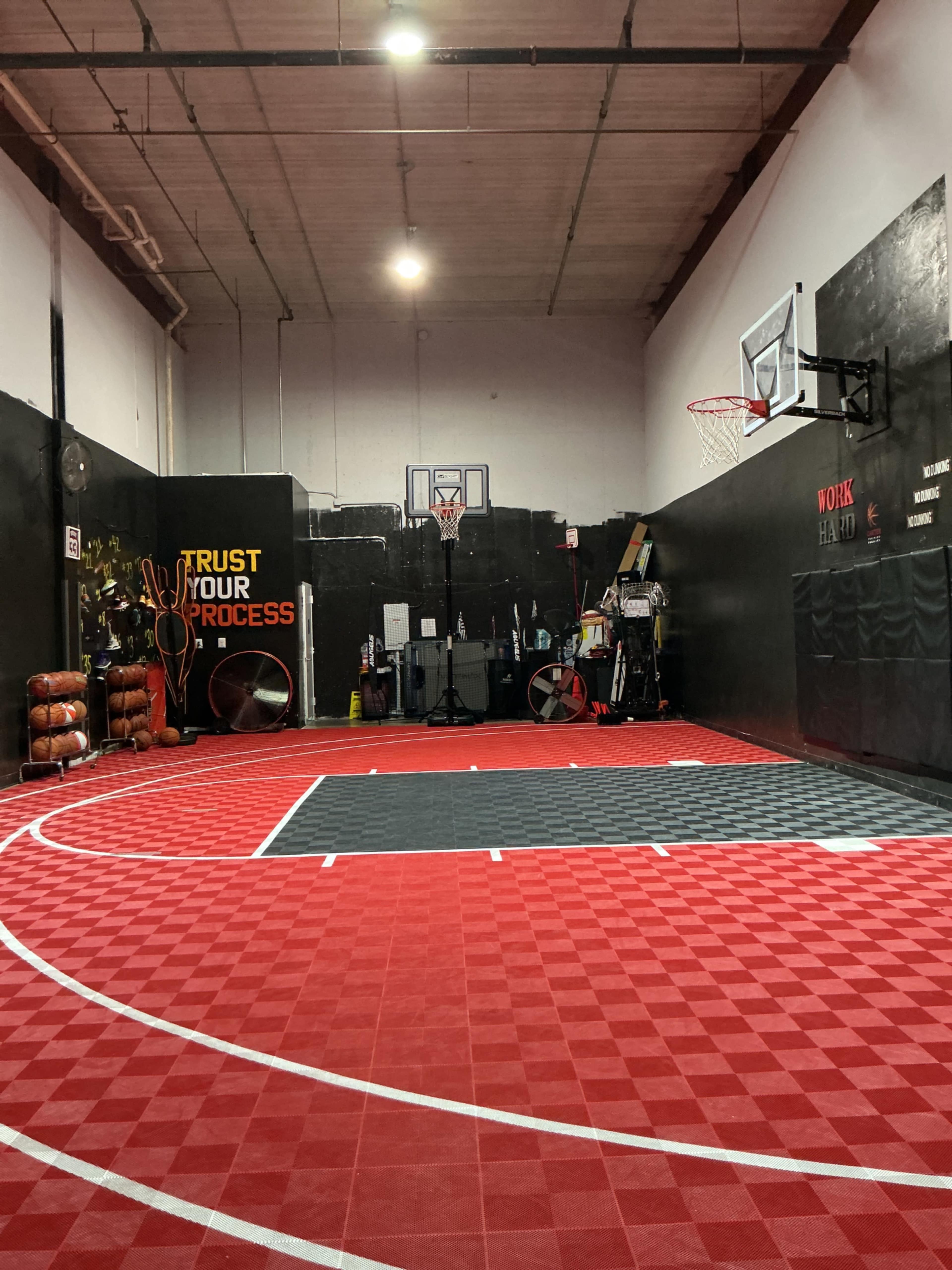 The image shows an indoor basketball court with a red and black checkered floor, featuring a basketball hoop at one end and various training equipment along the walls.
