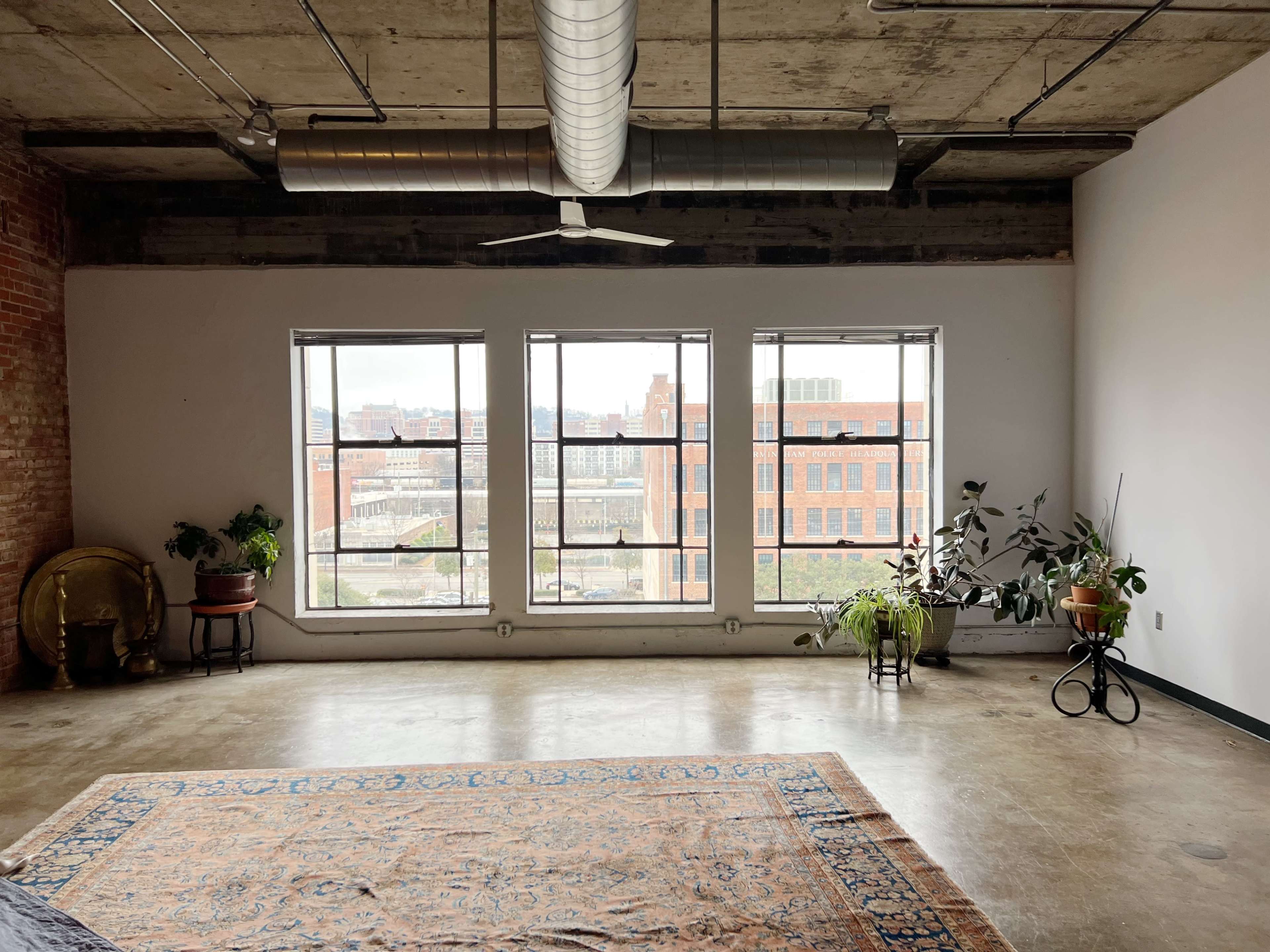 The image shows a spacious room with large windows overlooking an urban landscape, featuring a woven rug, potted plants, and industrial-style ceiling ductwork.