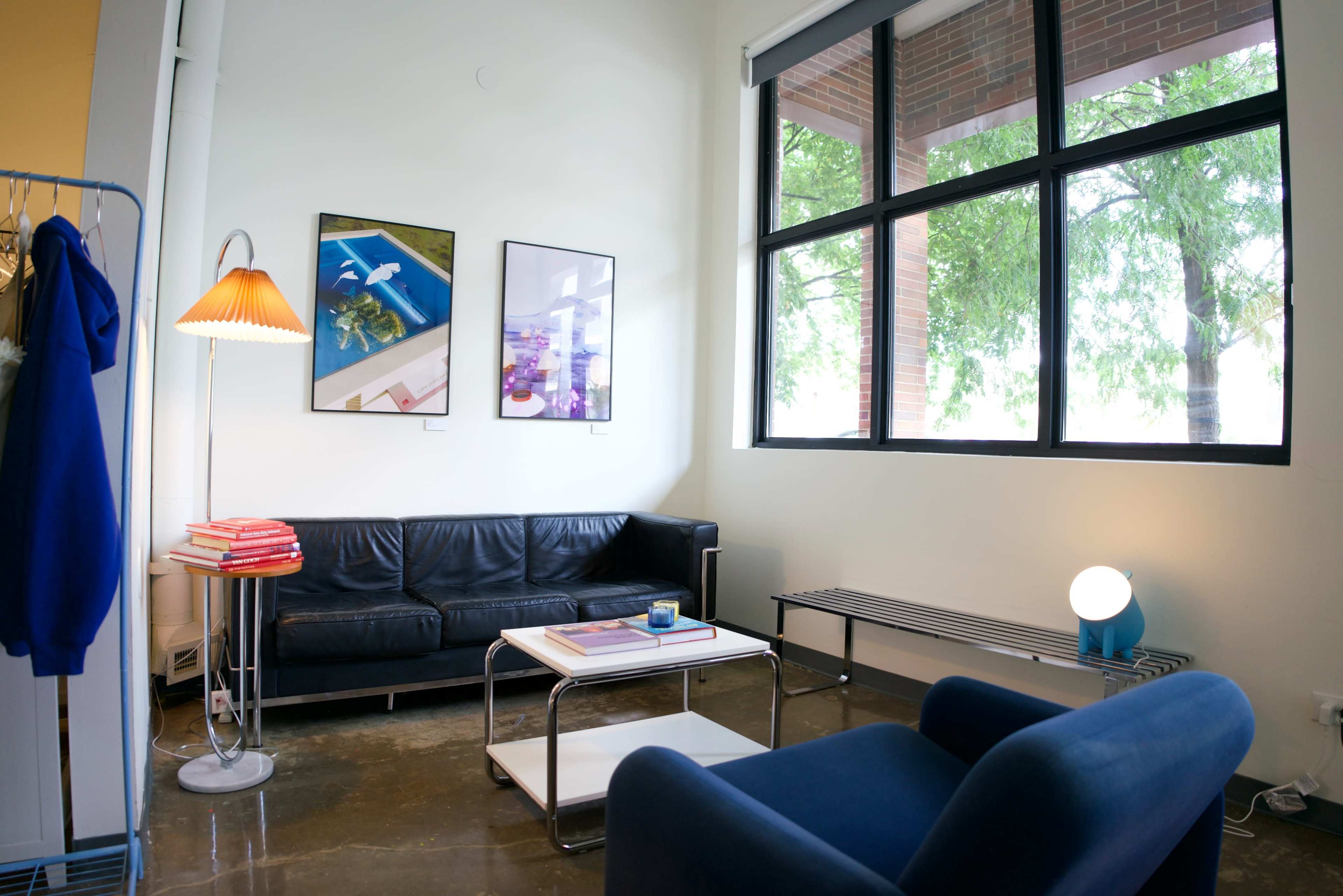 The image shows a modern living area with a black sofa, a blue armchair, a coffee table, and framed artwork on the walls, illuminated by natural light from large windows.