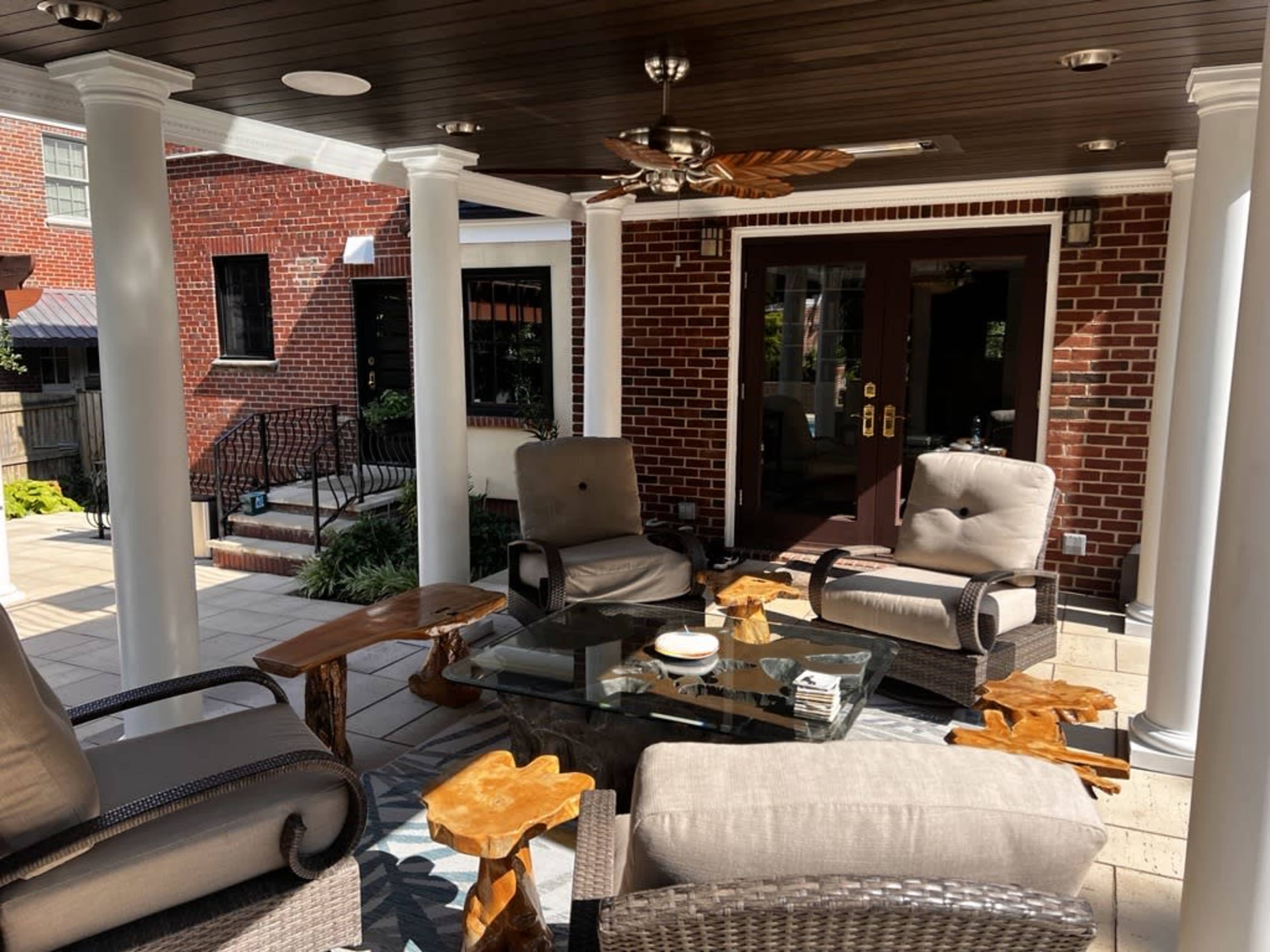 A covered outdoor patio with wicker furniture arranged around a glass table, surrounded by brick walls and columns.