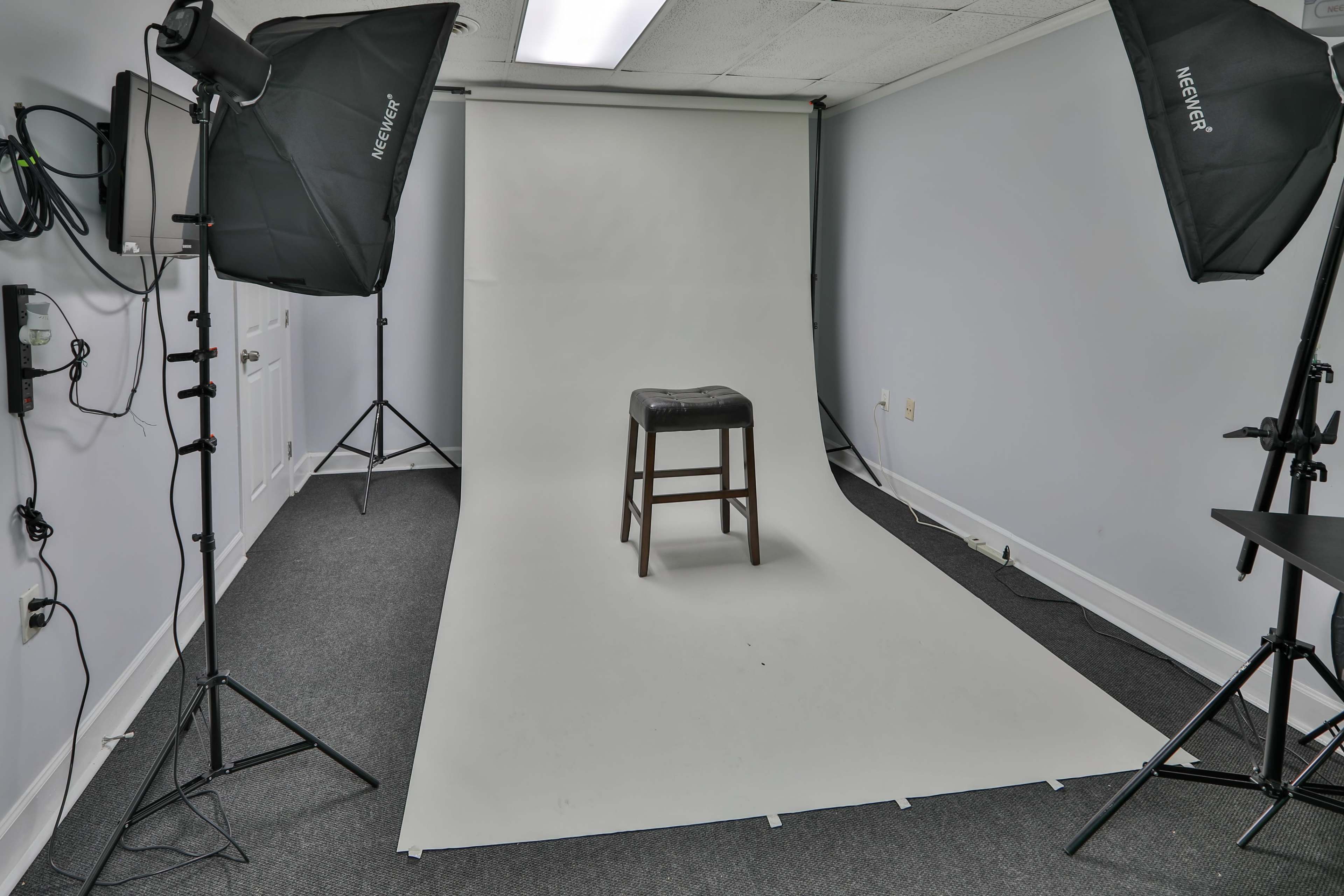 A photography studio features a bare white backdrop with a single wooden stool positioned in the center.