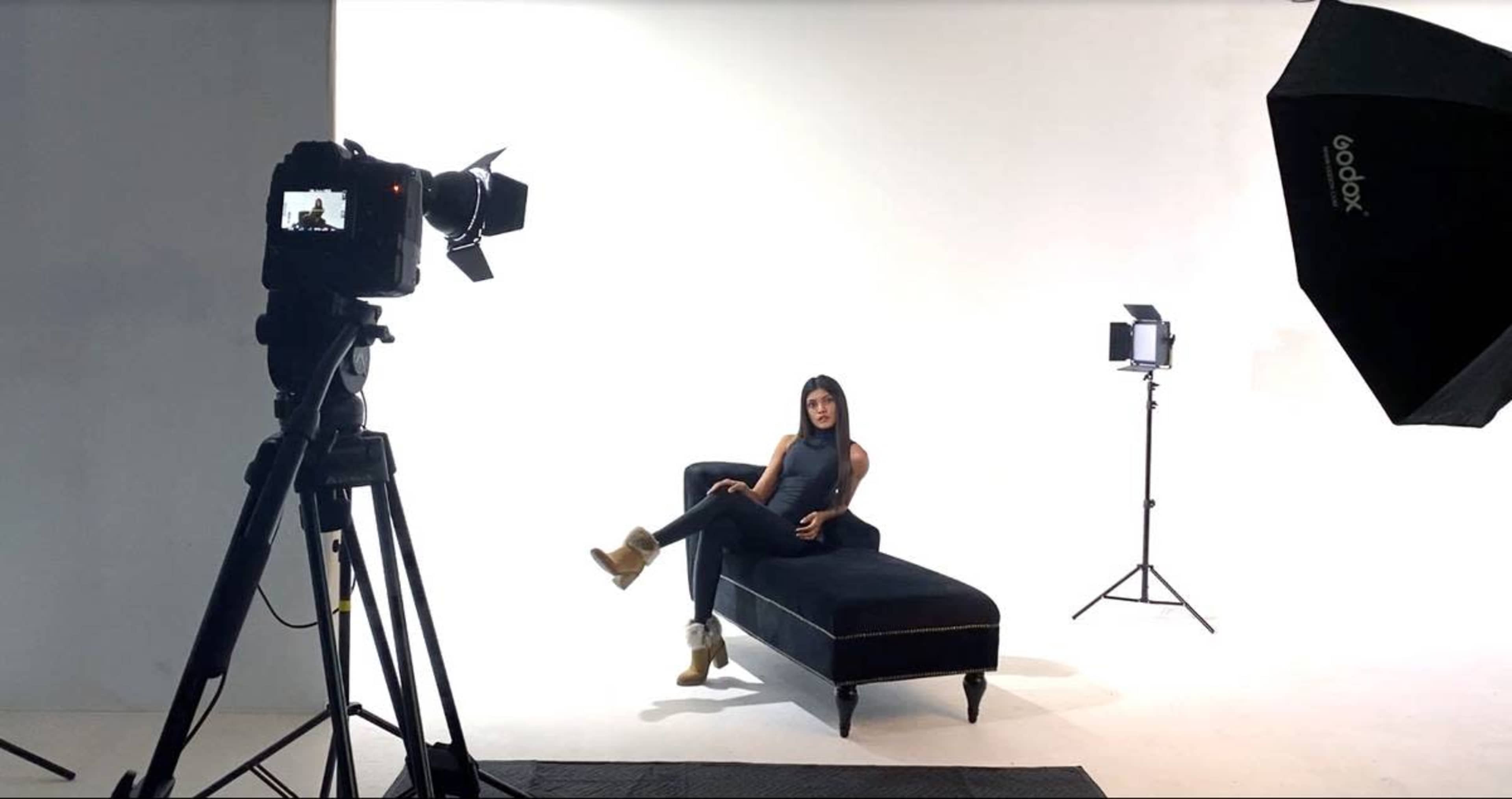 A woman sits on a black chaise lounge in a studio setting with a camera and lighting equipment positioned nearby.