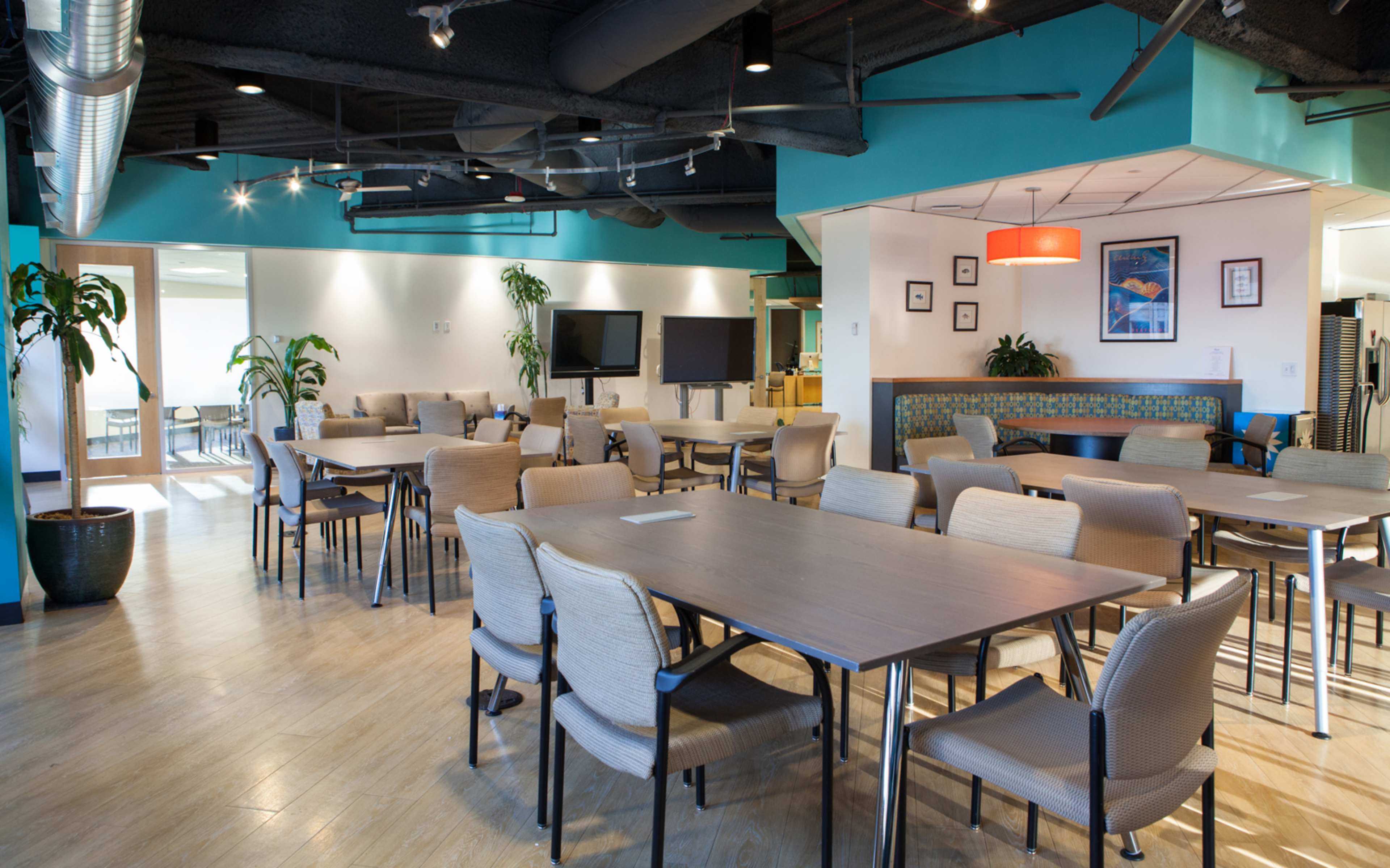 A modern conference room with multiple rectangular tables and chairs, surrounded by green plants and equipped with televisions on the walls.