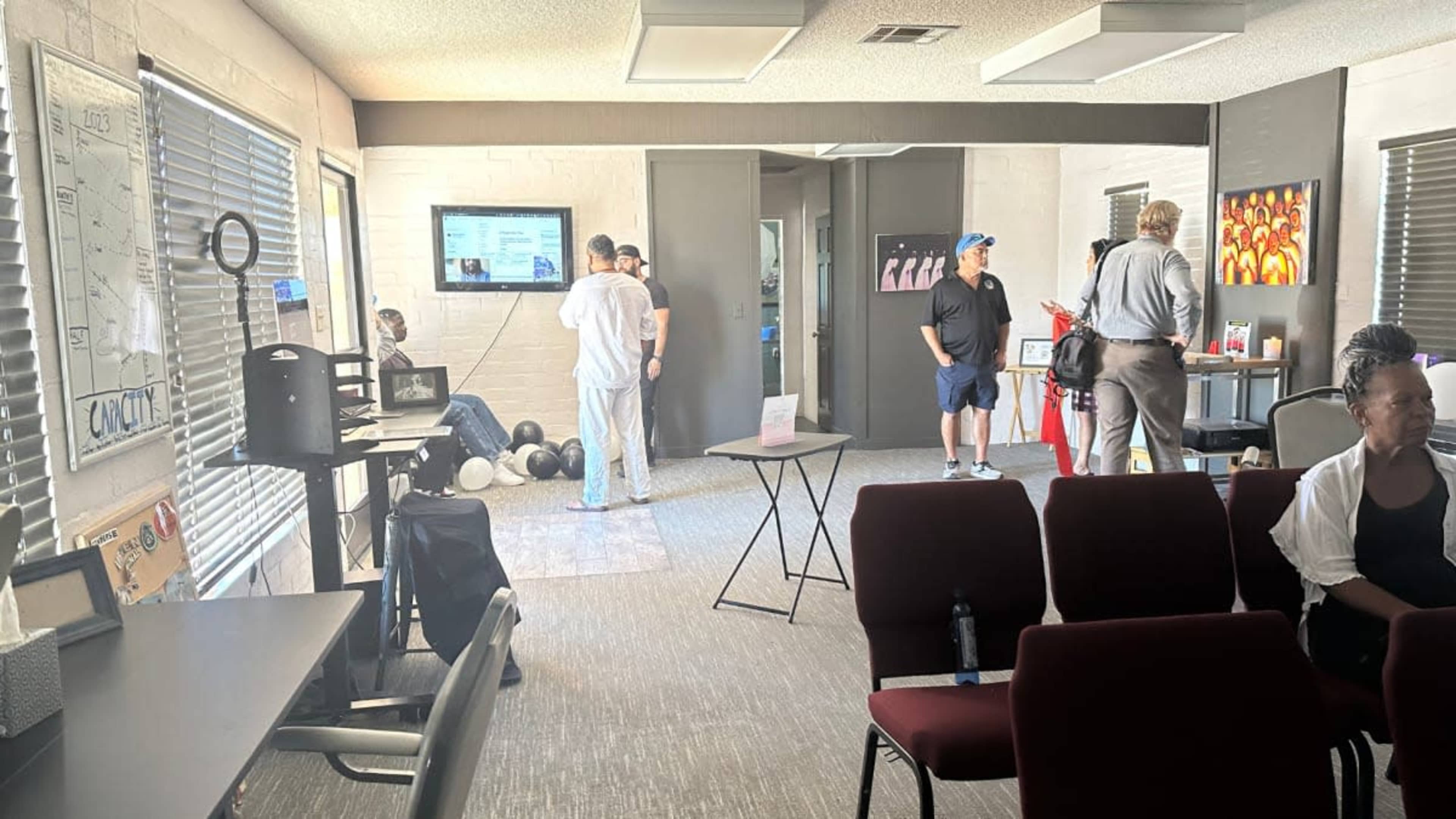 The image shows a spacious room with several people interacting, a large screen displaying information, and chairs arranged in a semi-circle.