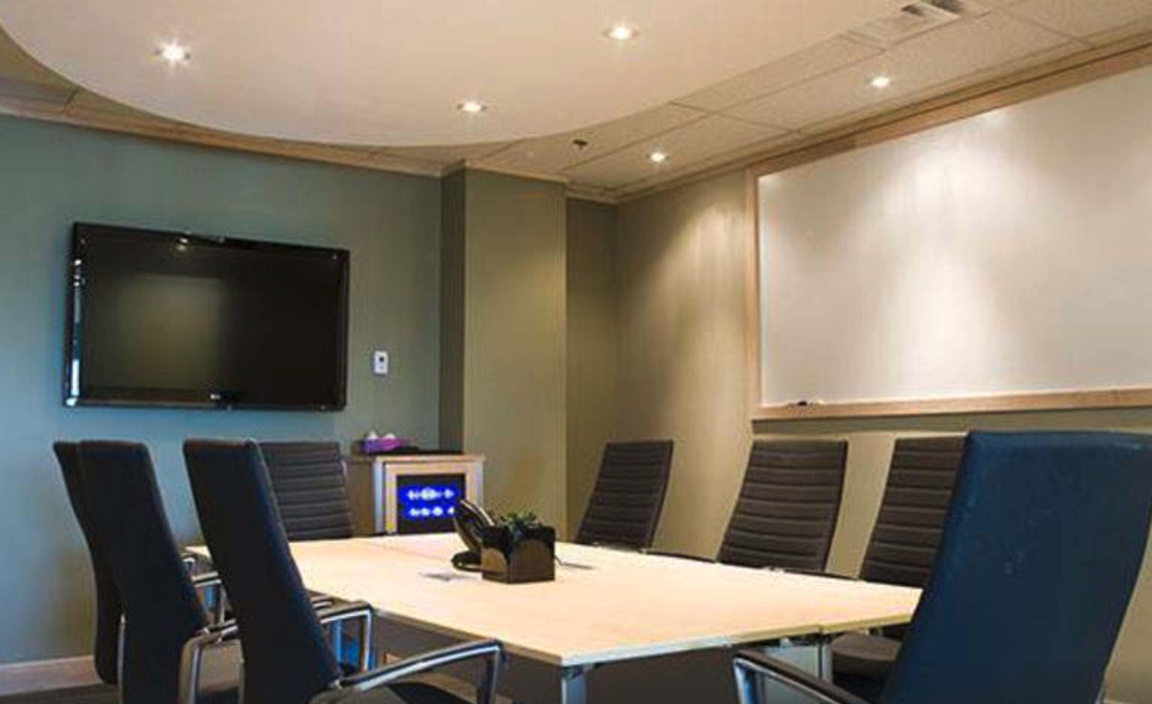 A modern conference room features a large table surrounded by black chairs, with a wall-mounted TV and a whiteboard in the background.