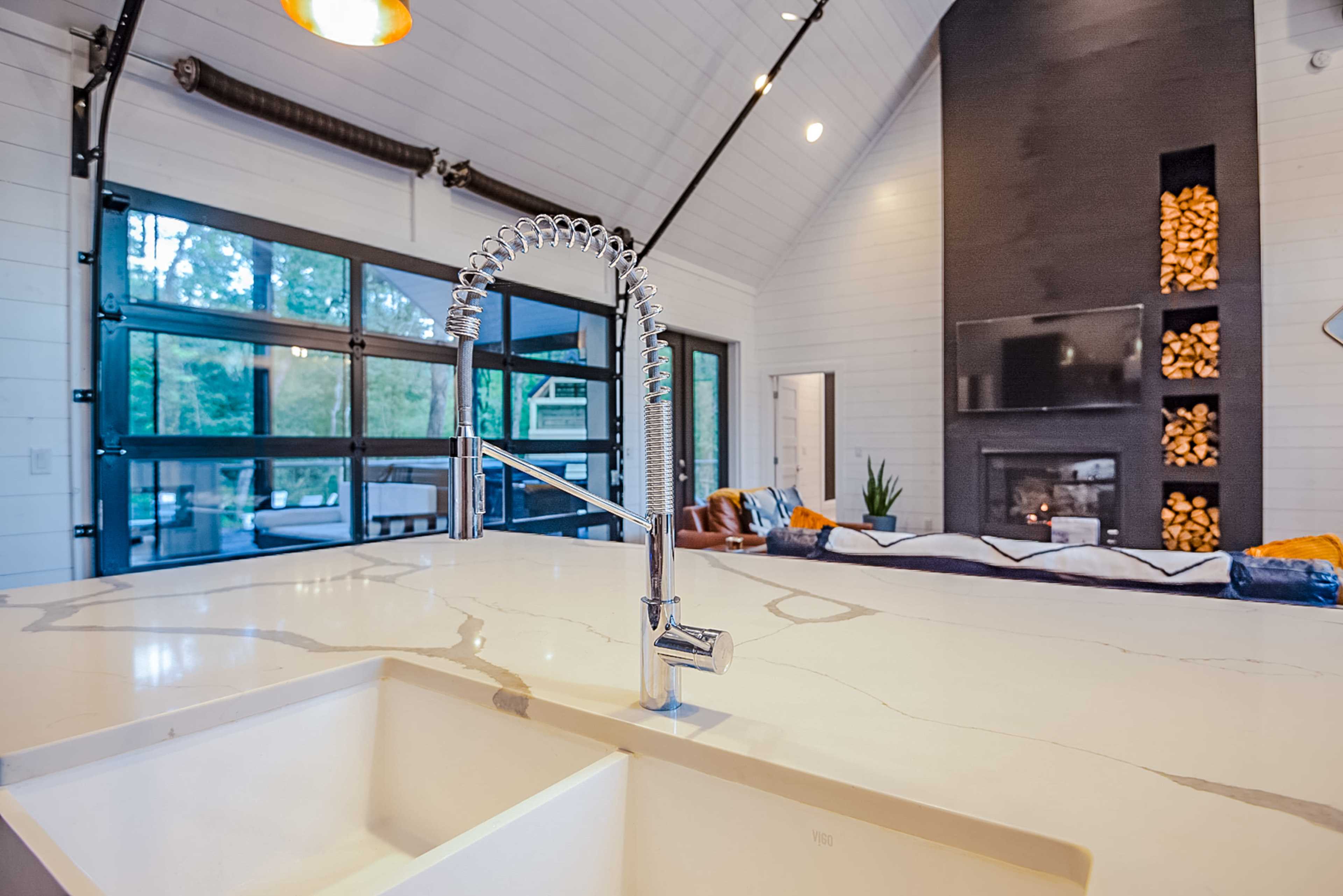 A modern kitchen with a chrome faucet and a large island, overlooking a living area with a fireplace and large glass doors.