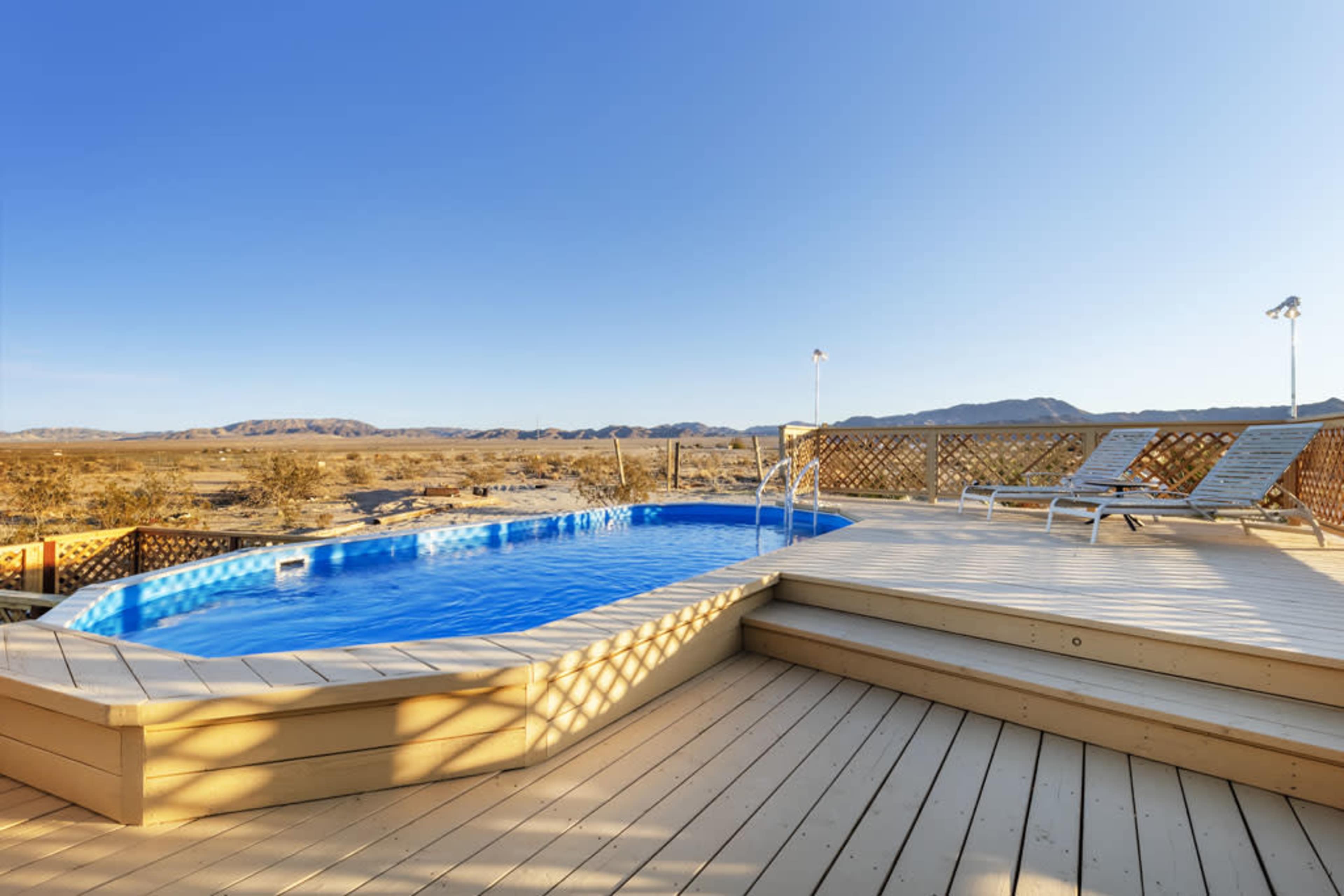 A rectangular swimming pool on a wooden deck with lounge chairs, surrounded by a desert landscape and mountains in the background.