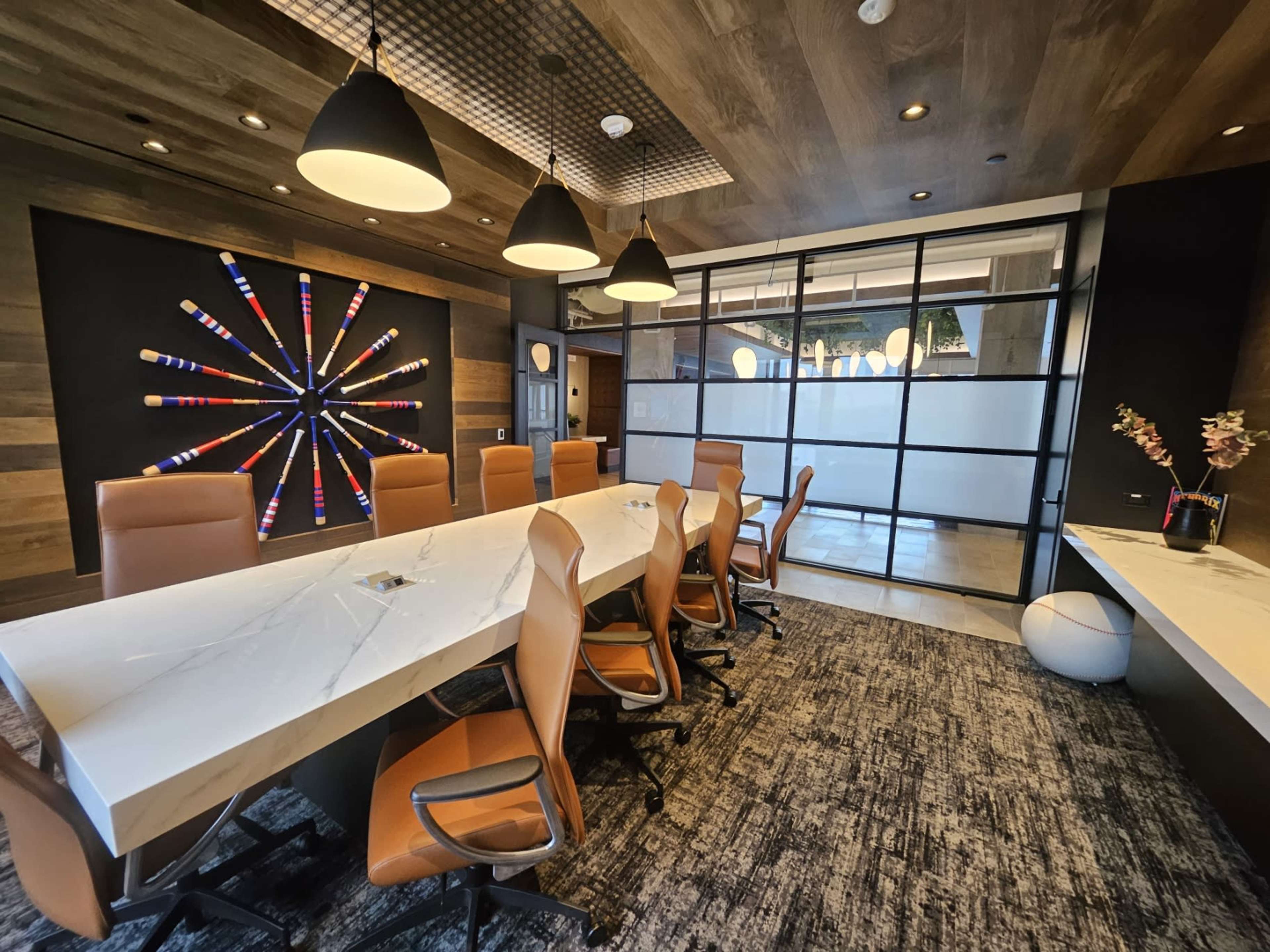 The image shows a modern conference room featuring a large white table surrounded by orange ergonomic chairs, with decorative artwork on the wall and a glass wall separating the room from an adjacent space.