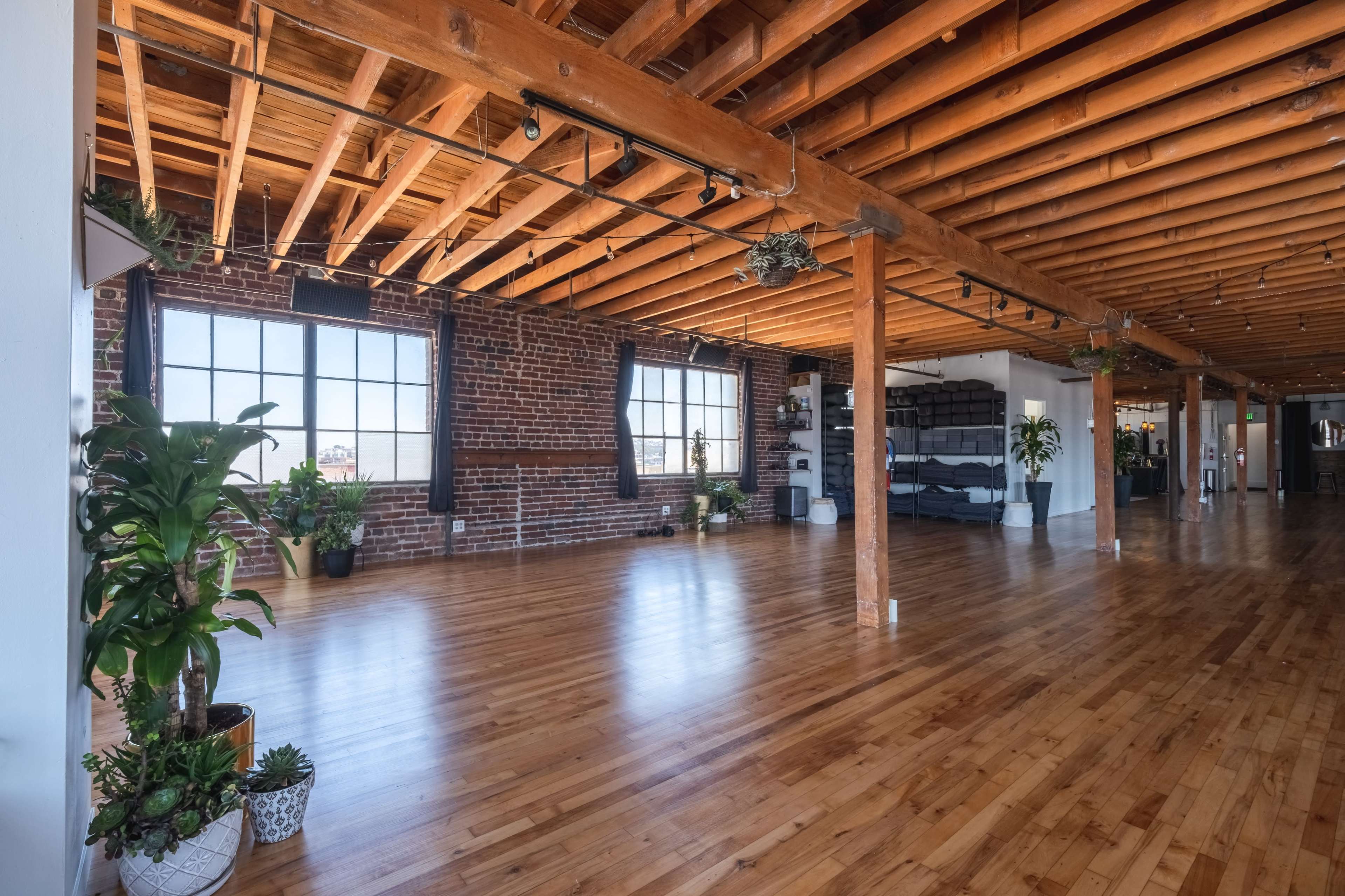 A spacious, bright room with wooden beams, hardwood floors, and large windows, decorated with various indoor plants.