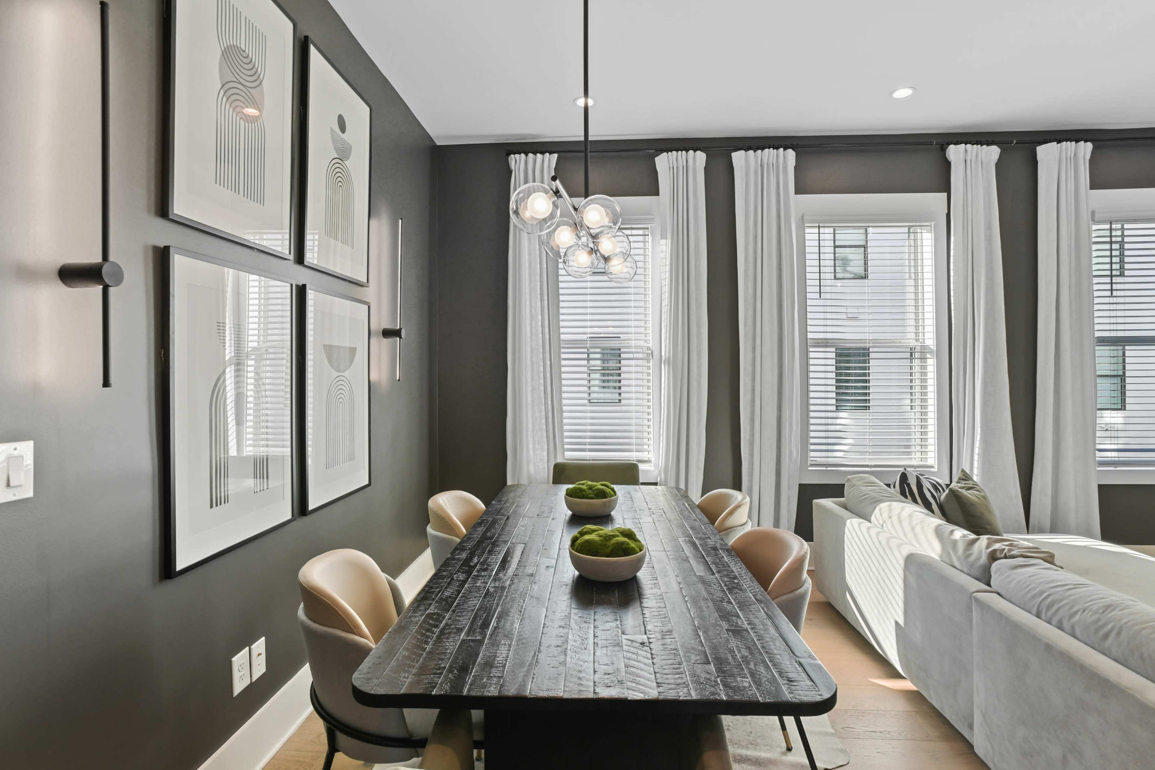 A dining area features a long black table surrounded by light-colored chairs, with art on the walls and large windows draped in white curtains.