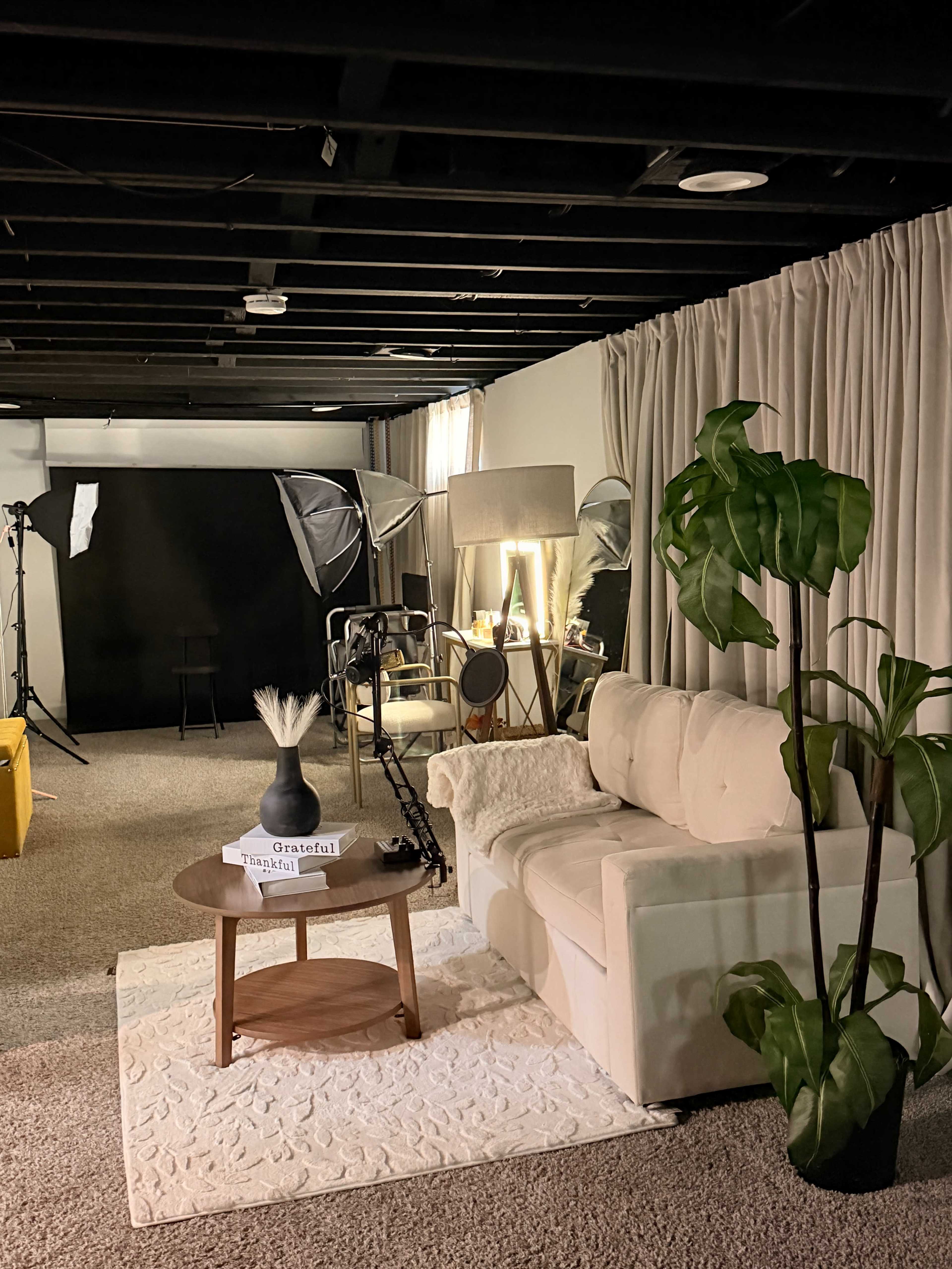 The image shows a dimly lit studio space featuring a white sofa, a circular wooden coffee table, a decorative plant, and various photography equipment in the background.