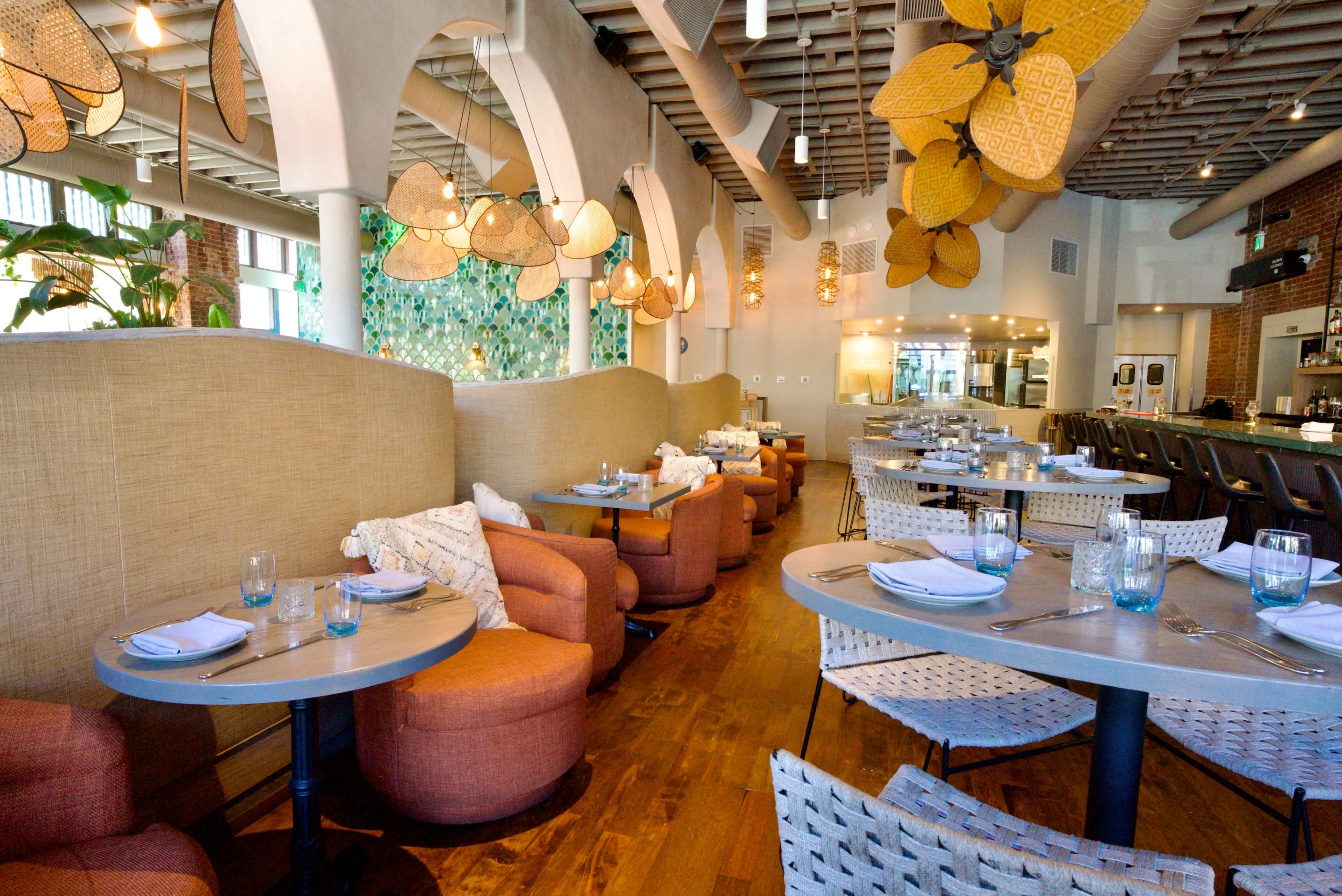 The image shows a brightly lit restaurant interior with circular tables, varied seating options, and decorative light fixtures overhead.