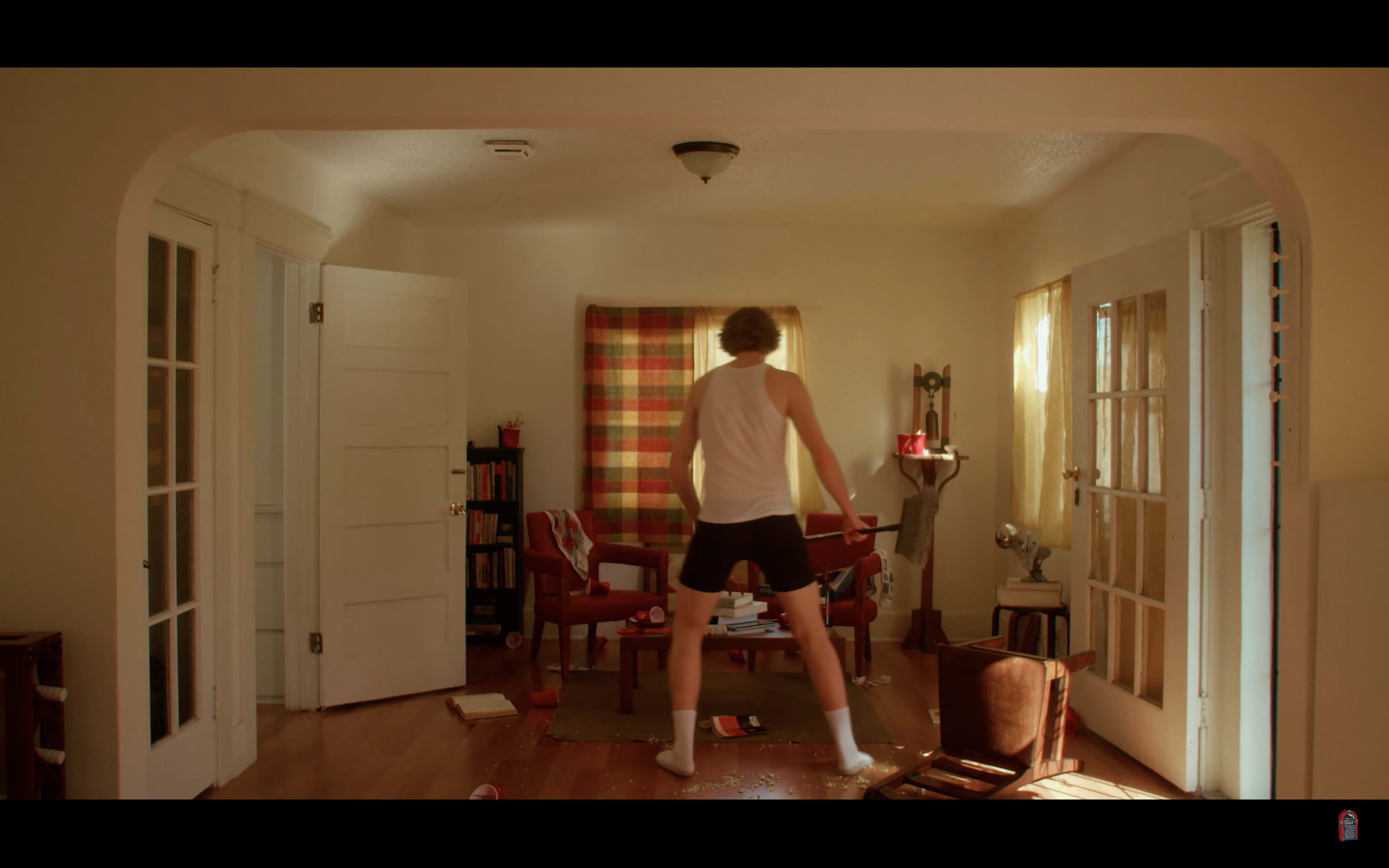 A man in a white tank top and shorts stands in a disordered living room with scattered books and furniture.