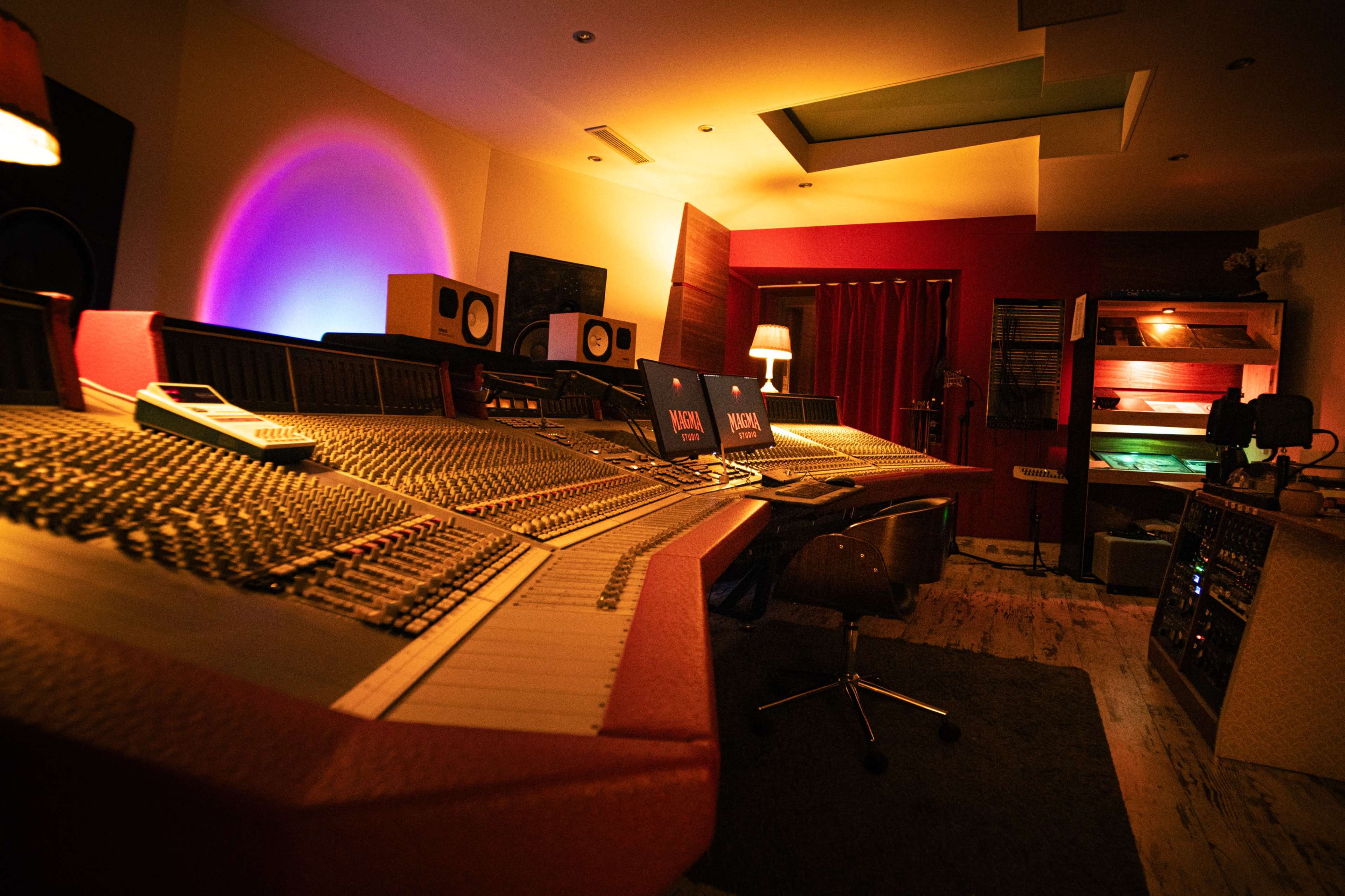 The image depicts a recording studio with a large mixing console, computer screens, and various audio equipment arranged around a spacious room with ambient lighting.