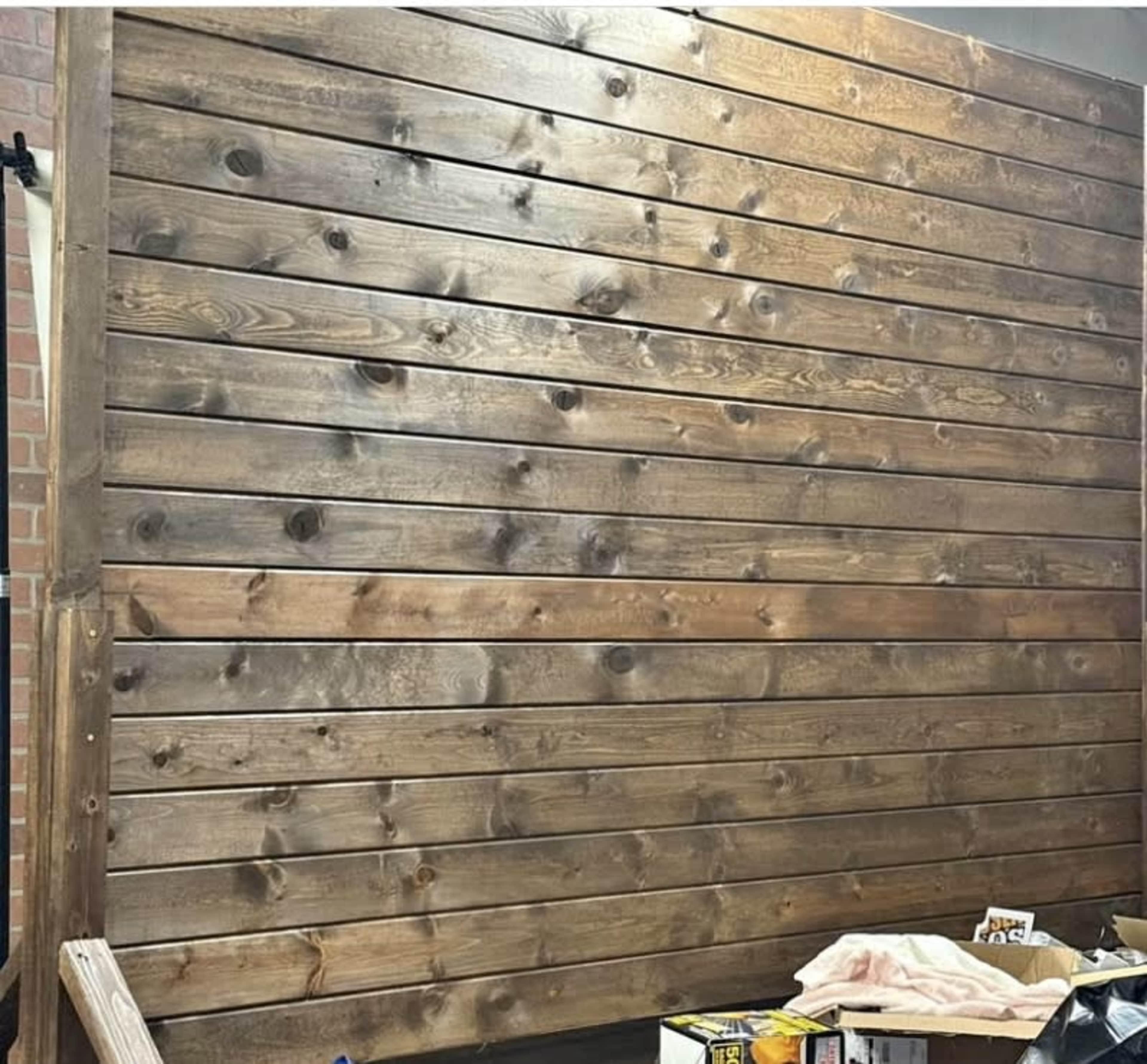 The image features a vertical wooden wall with horizontal planks arranged in a stacked pattern, surrounded by various materials on the floor.
