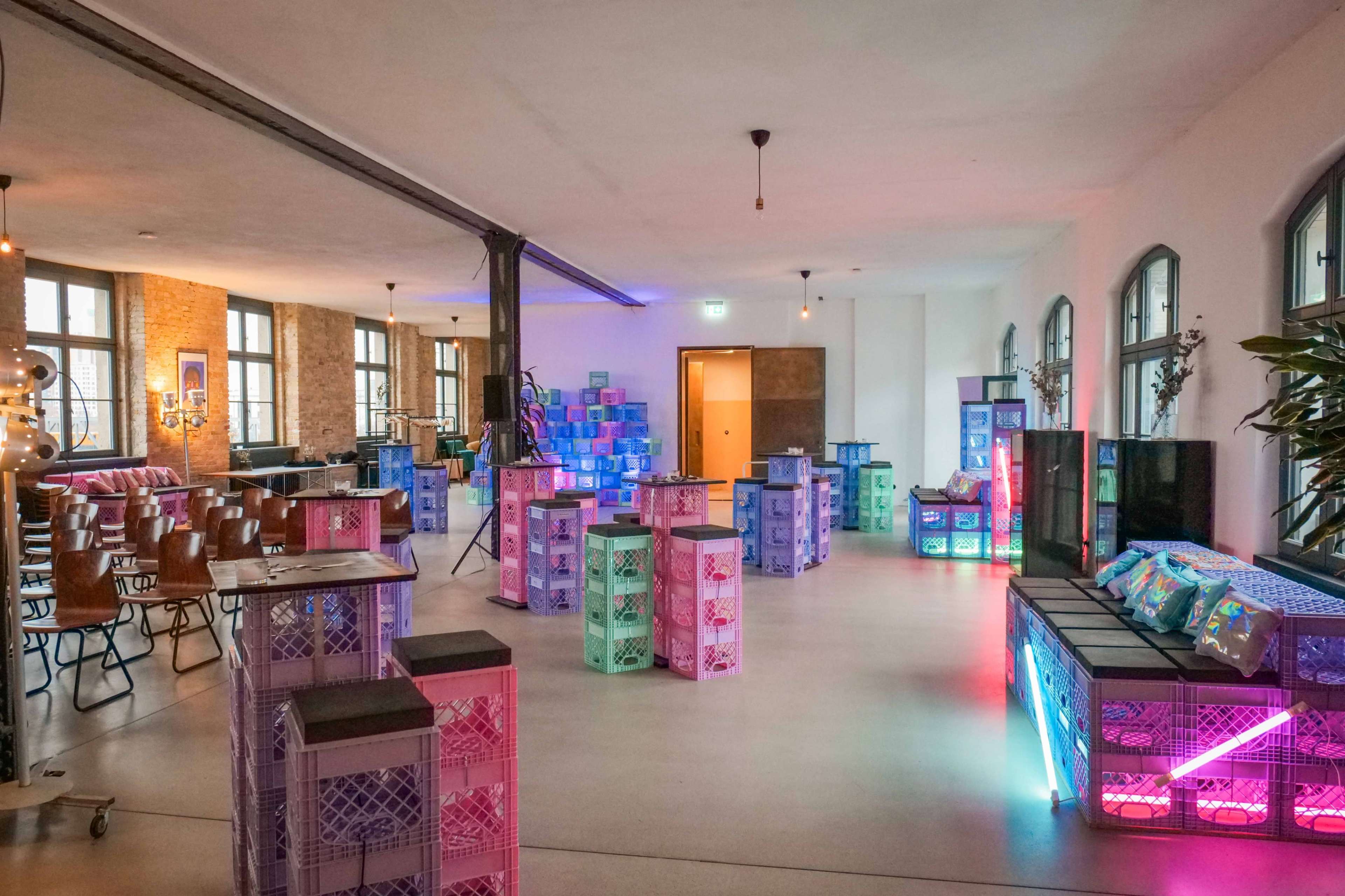 The image shows a spacious room furnished with colorful stacked crates serving as tables and seating, illuminated by various lights.